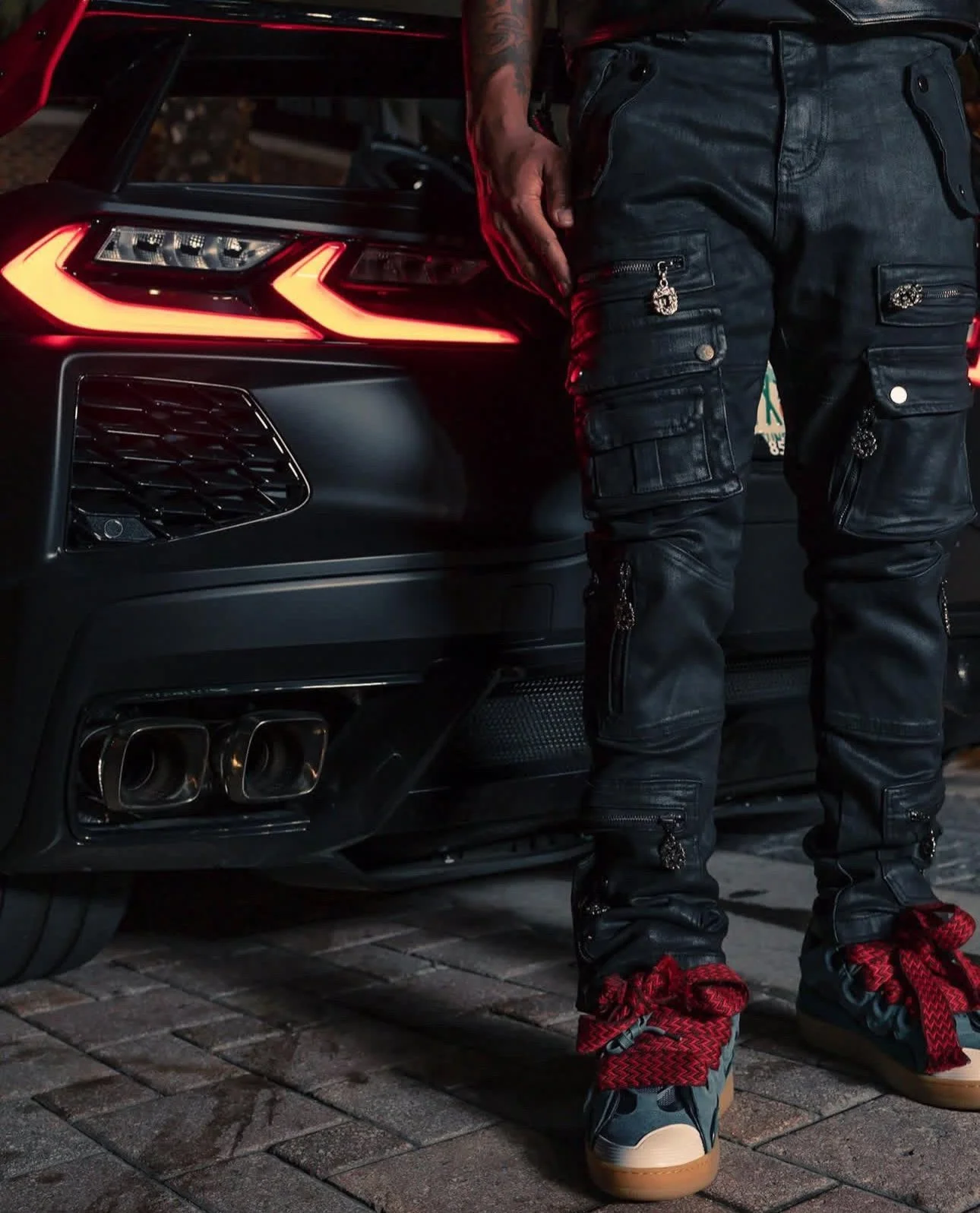 Person standing beside a black car at night, wearing black cargo pants with zippers and patches, and red and black sneakers.