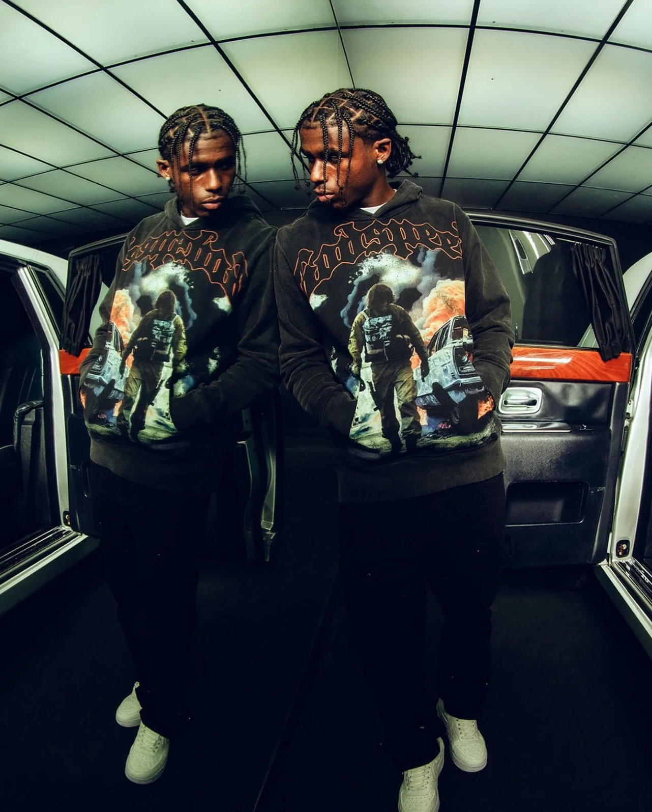 Two young men standing in front of a car with open doors, wearing matching black hoodies featuring a graphic of a person walking through smoke with a skyline, inside a room with reflective ceiling panels.