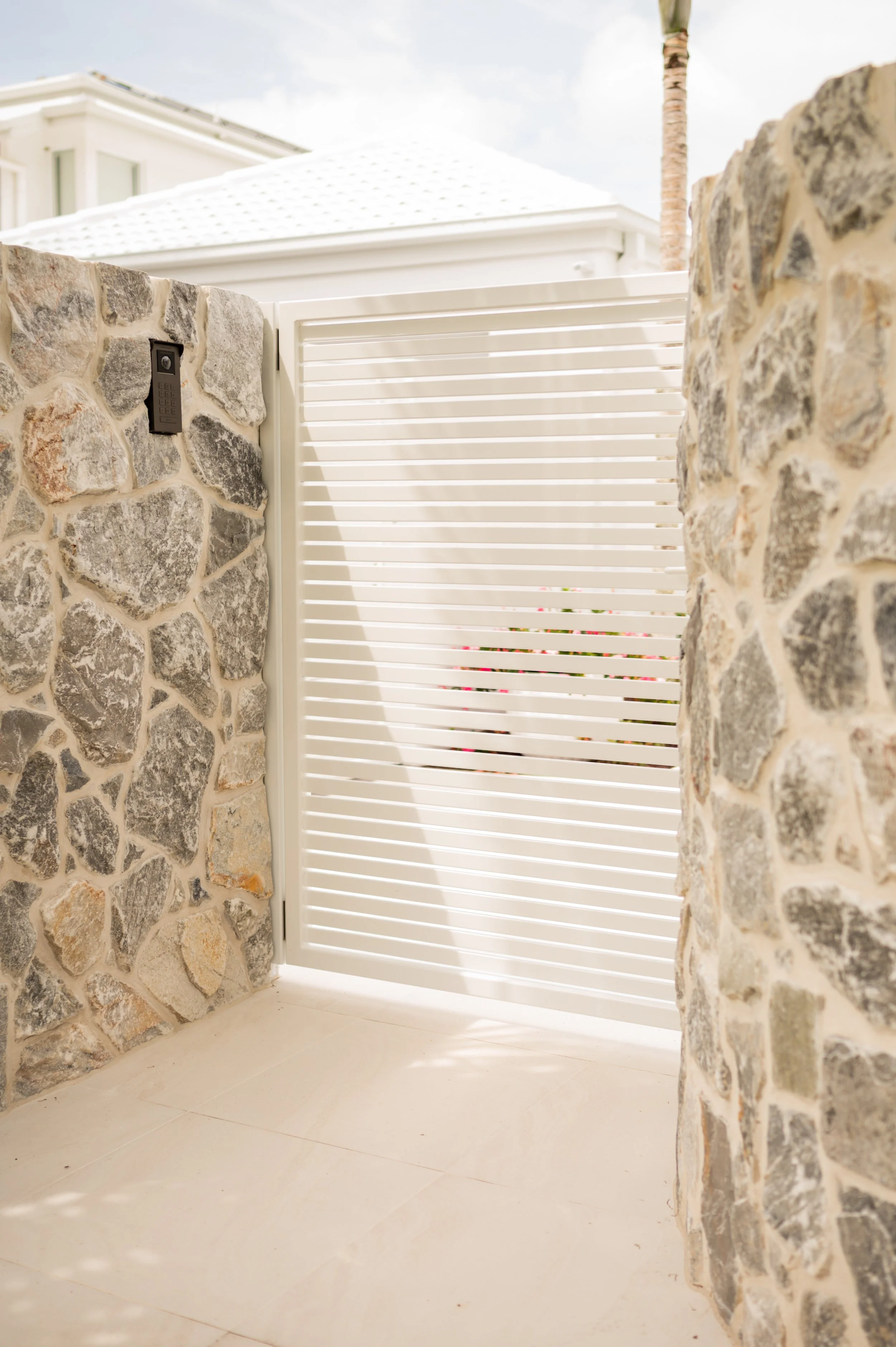 Stone wall, white slatted gate, intercom, and part of a white building with a roof in the background.