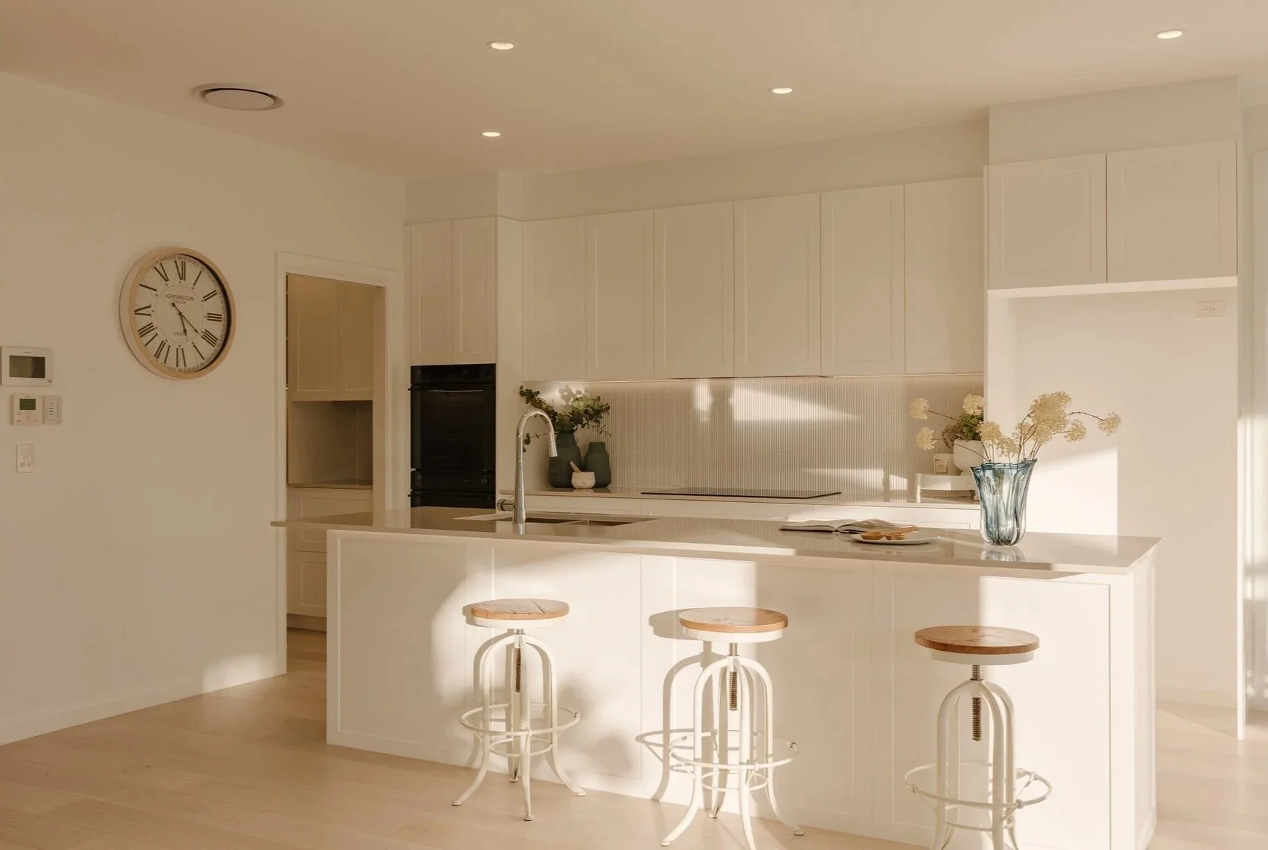 Bright, minimalist kitchen with white cabinetry, a central island with a sink, three wooden and metal barstools, a large clock on the wall, and decorative vases with flowers on the counter and island. Sunlight streams in.