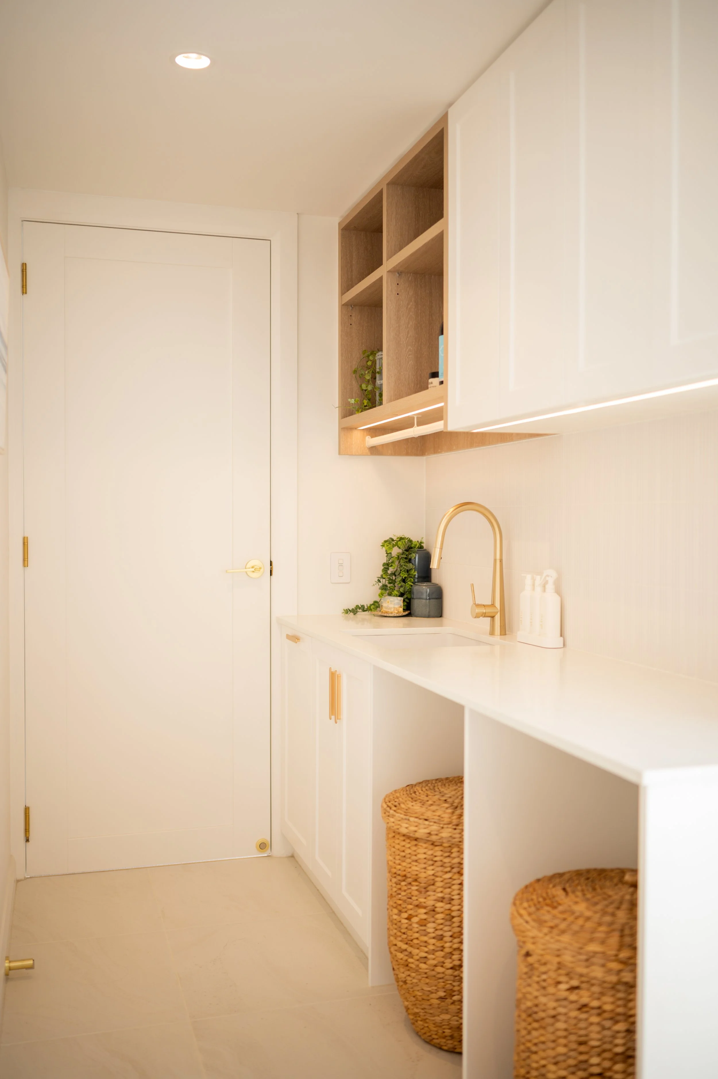 White kitchen with gold faucet, open shelving, beige woven baskets, and small potted plants.