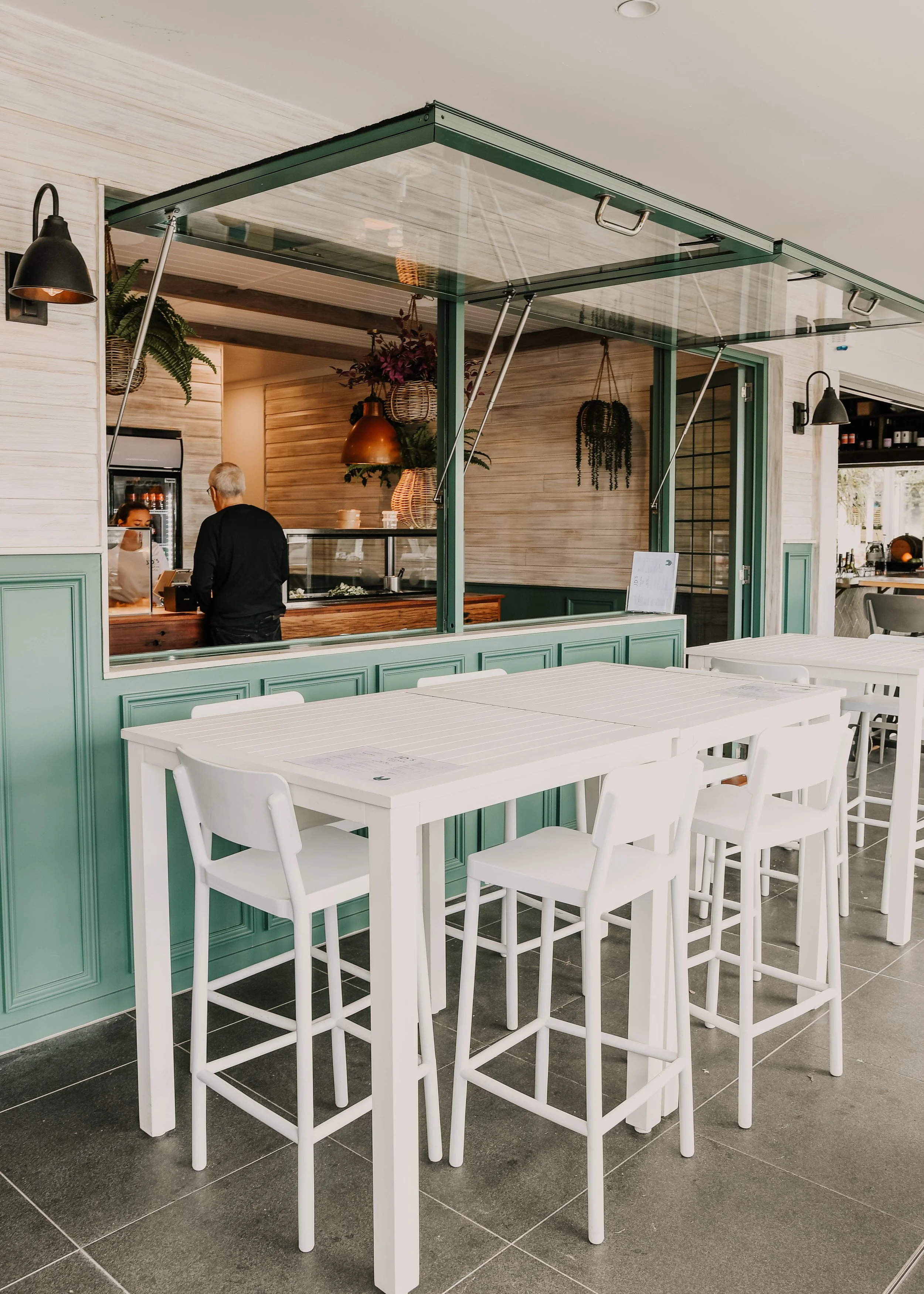 Empty white tables and chairs outside a restaurant with an open window, interior with wooden wall, plants, hanging lights, and a person ordering food.