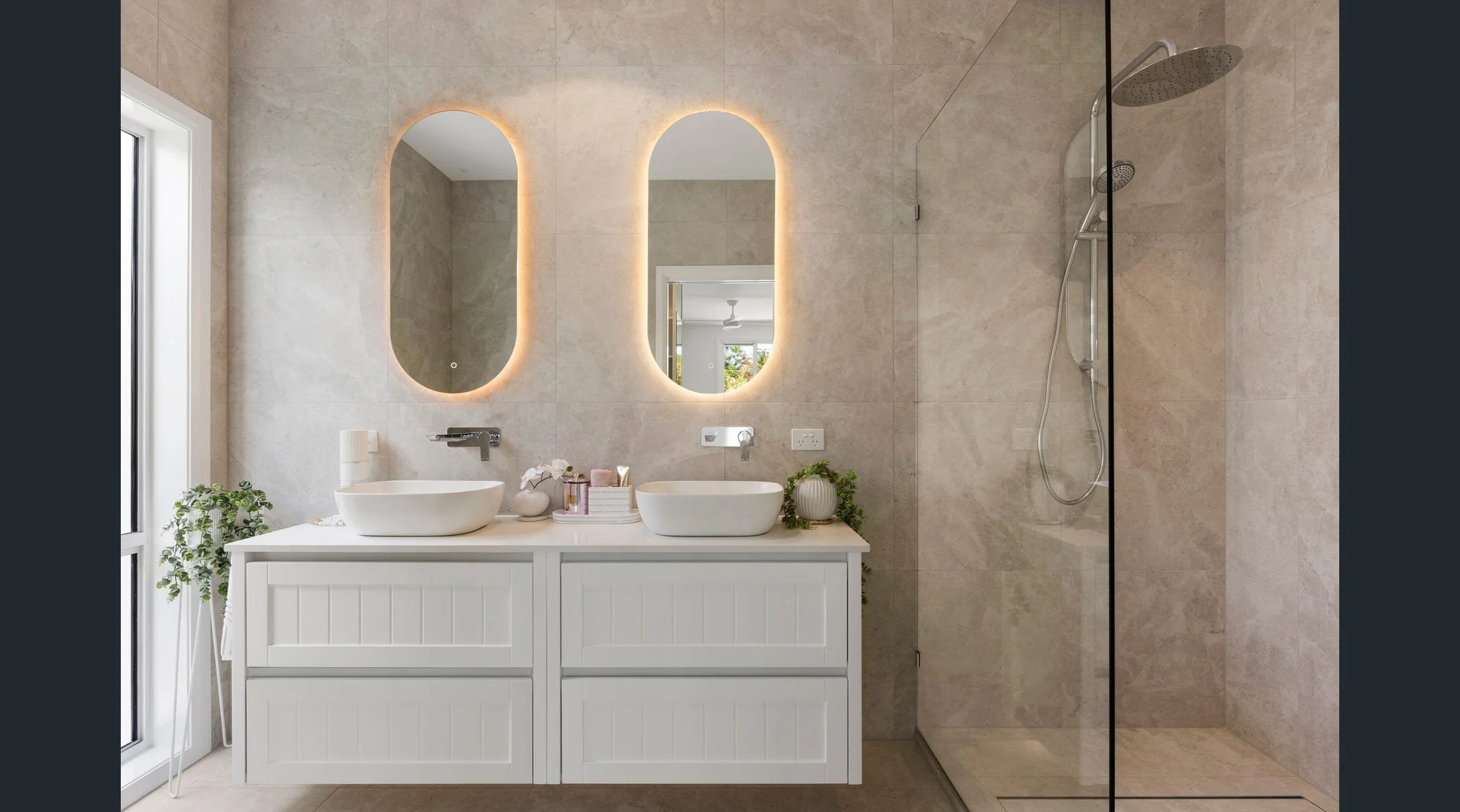 Modern bathroom with dual white vessel sinks on a white cabinet, large backlit mirrors, a walk-in shower with a rain showerhead, and natural light from a window.