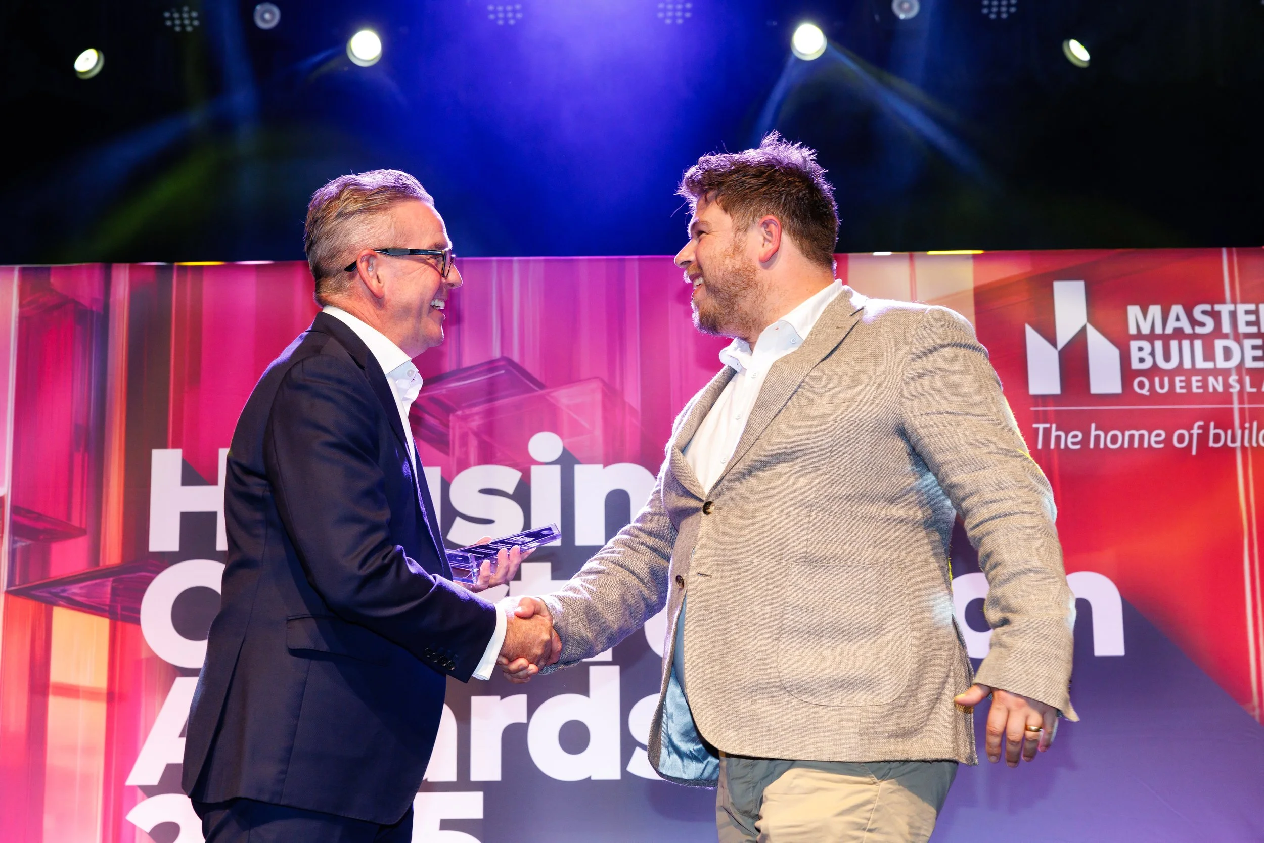 Two men in suits shaking hands on stage at an award ceremony, with a colorful backdrop displaying the words 'Housing Industry' and 'Queensland Master Builders'.