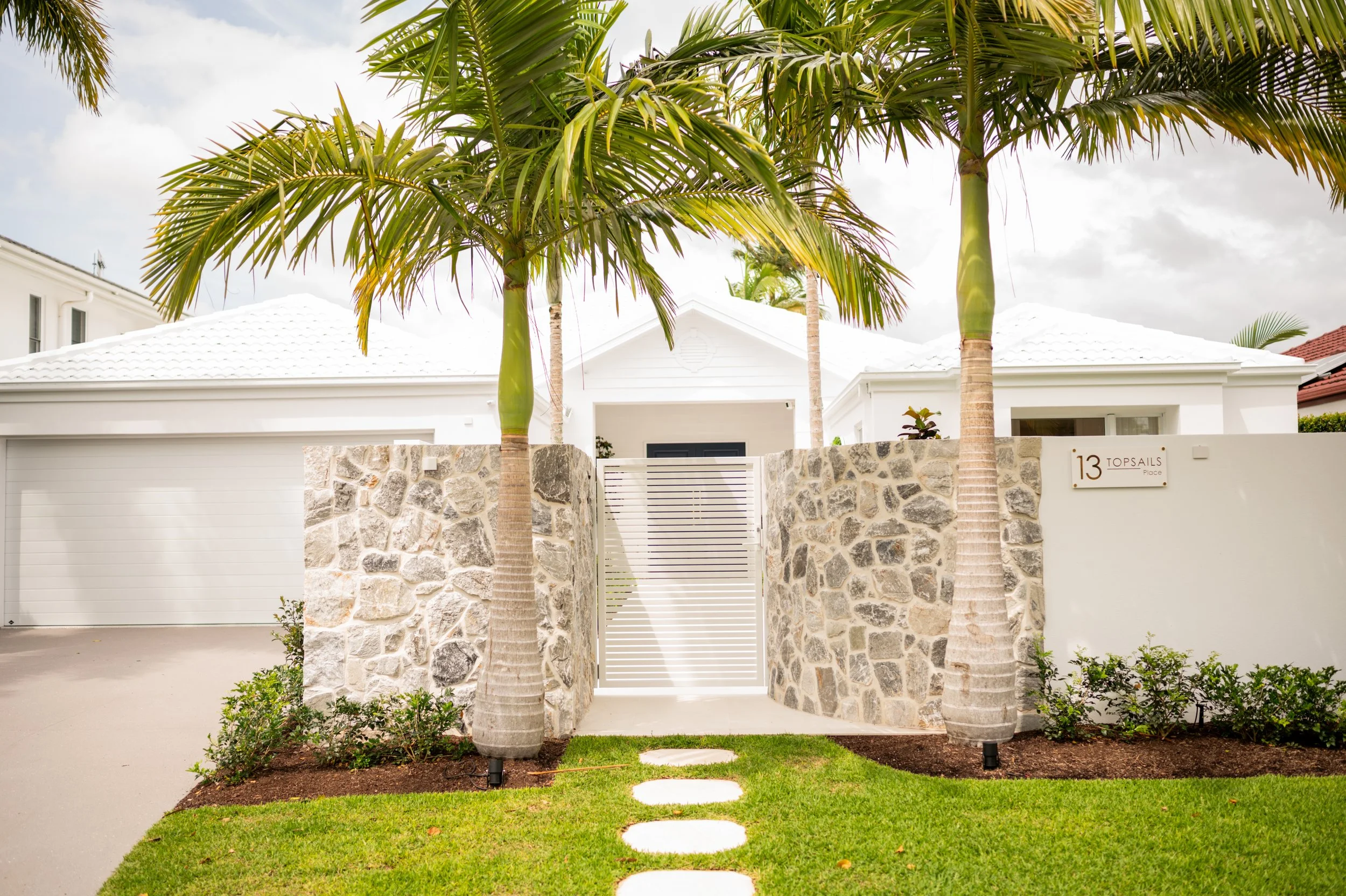 Modern white house with a stone wall, white gate, palm trees, and a well-manicured lawn.