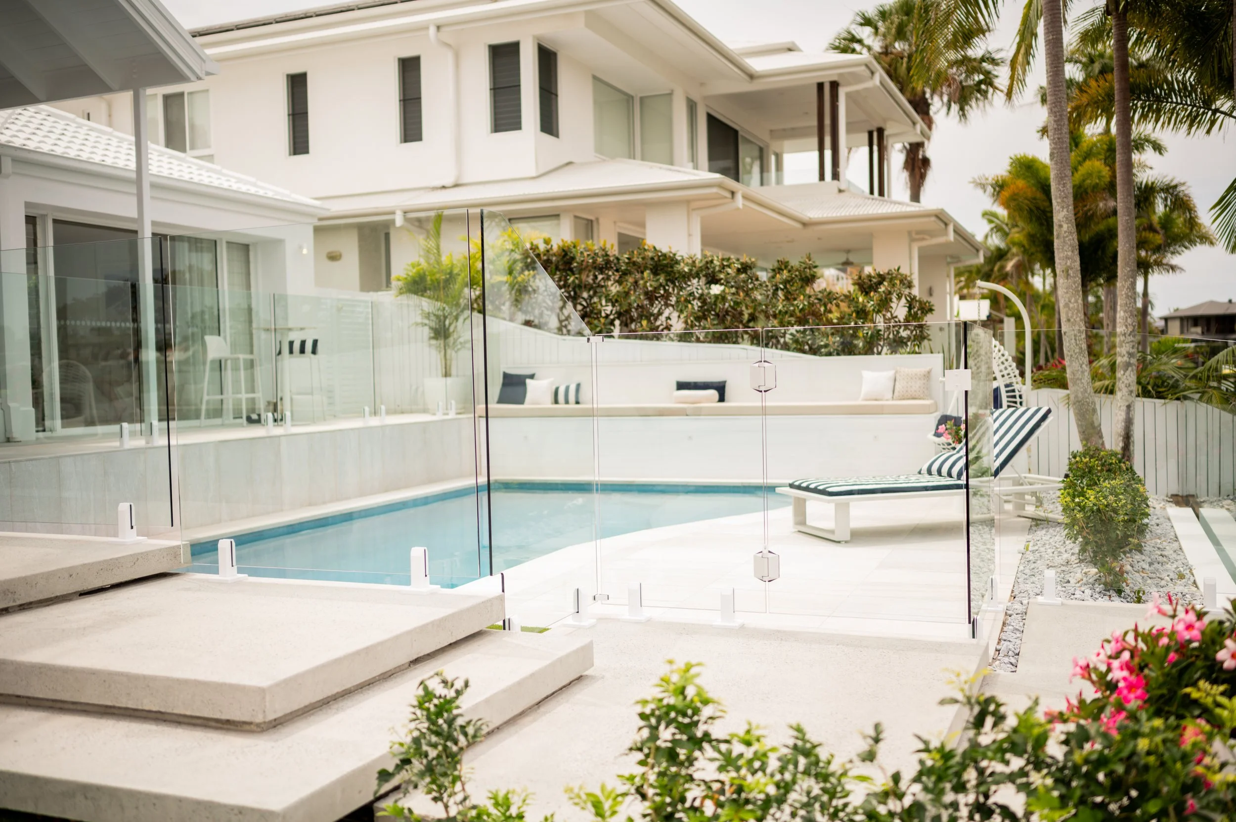 Luxurious backyard with a small swimming pool surrounded by a glass fence, a reclining lounge chair with striped cushion, lush palm trees, and a modern white house with multiple balconies in the background.