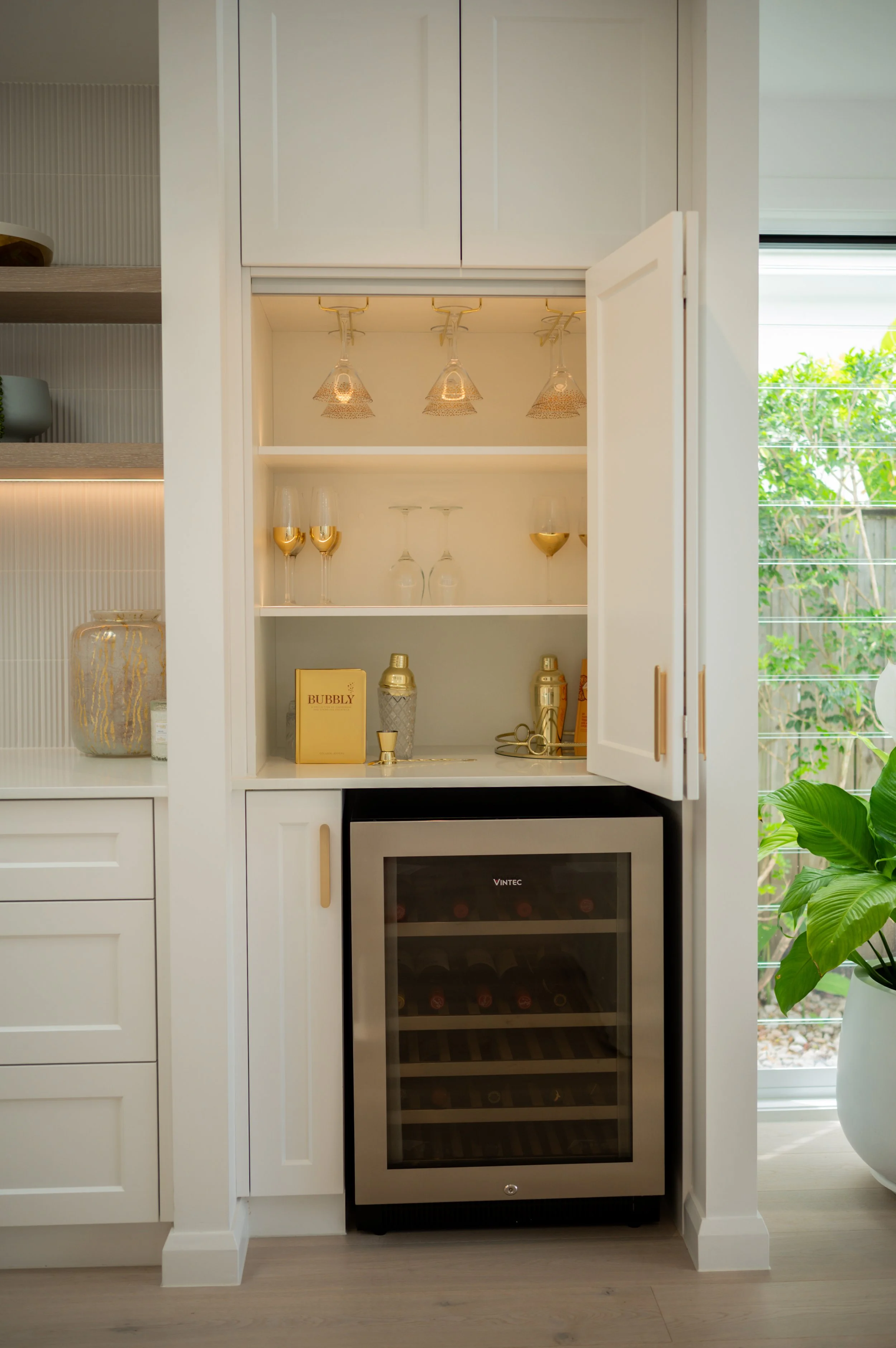 Built-in bar area with glassware, gold accents, and a wine cooler in a modern kitchen.