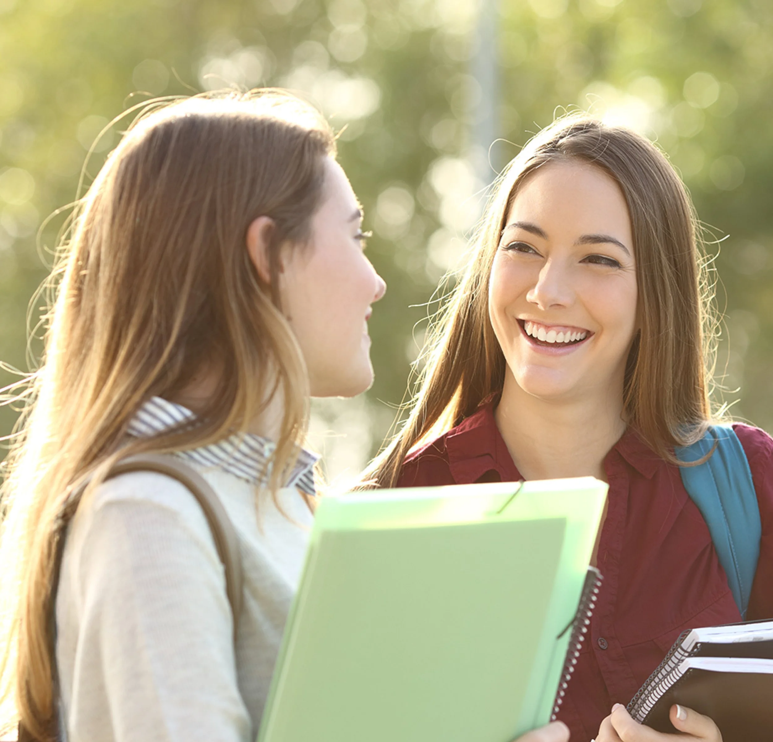 Two female students smile at one another while holding books outside