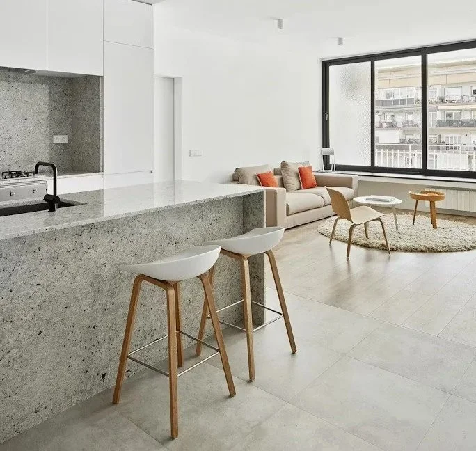Modern open-plan living room and kitchen with a large window, light-colored furniture, and a terrazzo kitchen island with two white bar stools.
