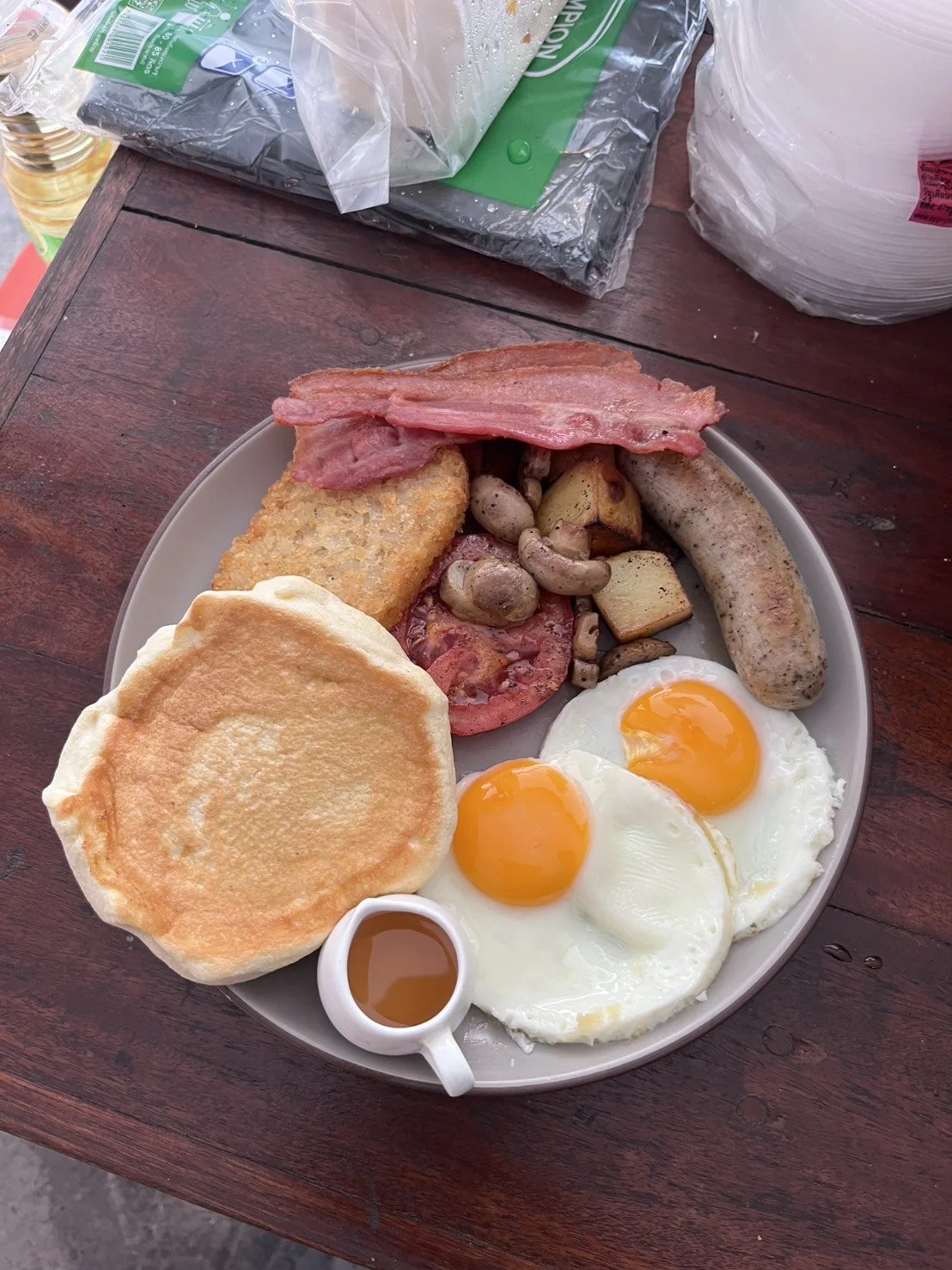 A plate of breakfast food including sunny side up eggs, fried bread, bacon, sausage, sautéed mushrooms and potatoes, and a small container of syrup, on a wooden table.