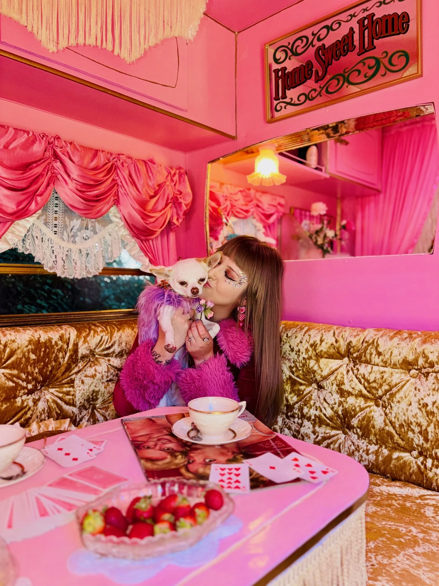 A woman with long brown hair, tattoos on her arms, and wearing a pink shirt, smiling while holding a small brown and white puppy dressed in a tuxedo with a red bow tie.