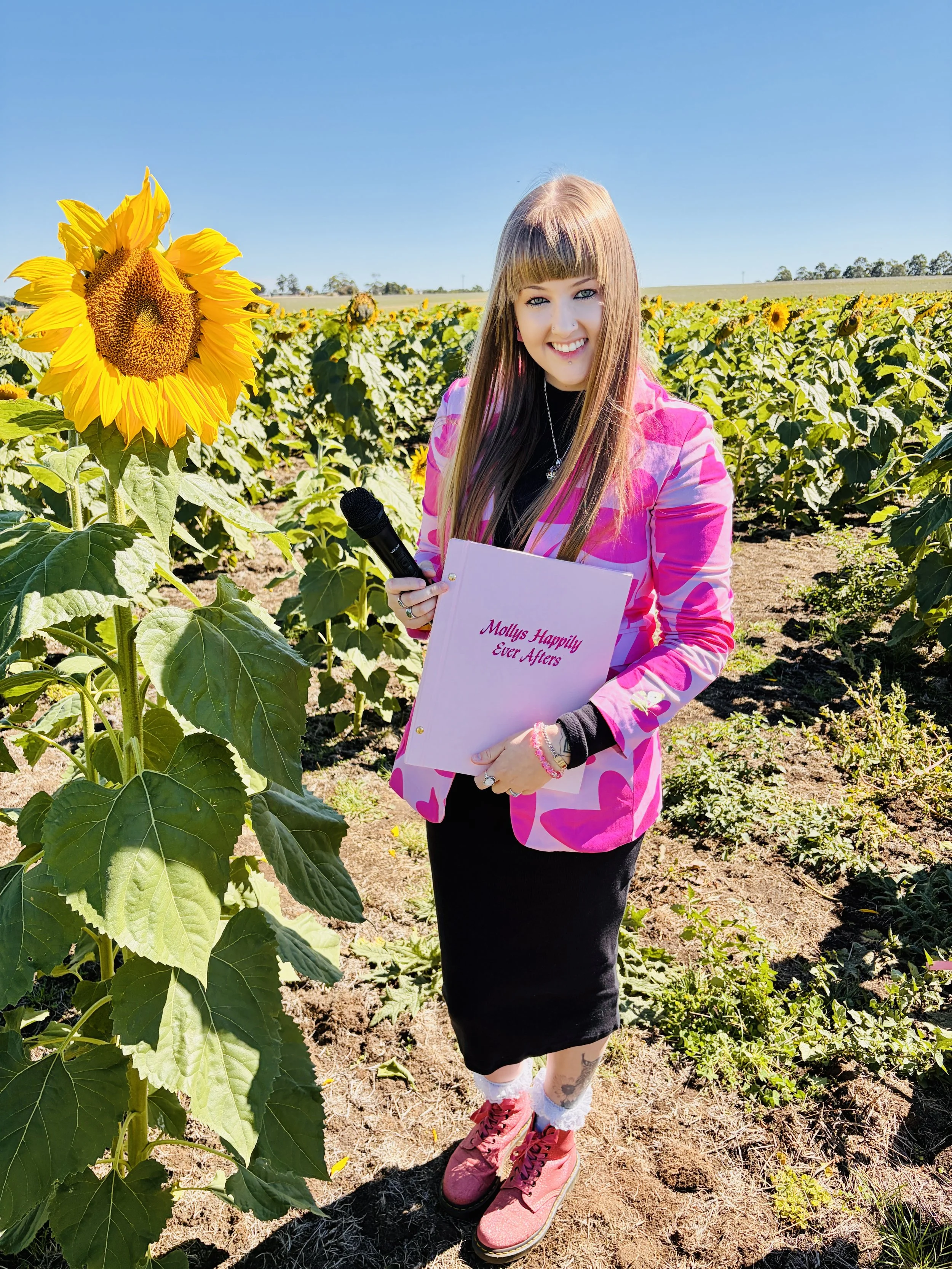 A woman with long brown hair, tattoos on her arms, and wearing a pink shirt, smiling while holding a small brown and white puppy dressed in a tuxedo with a red bow tie.