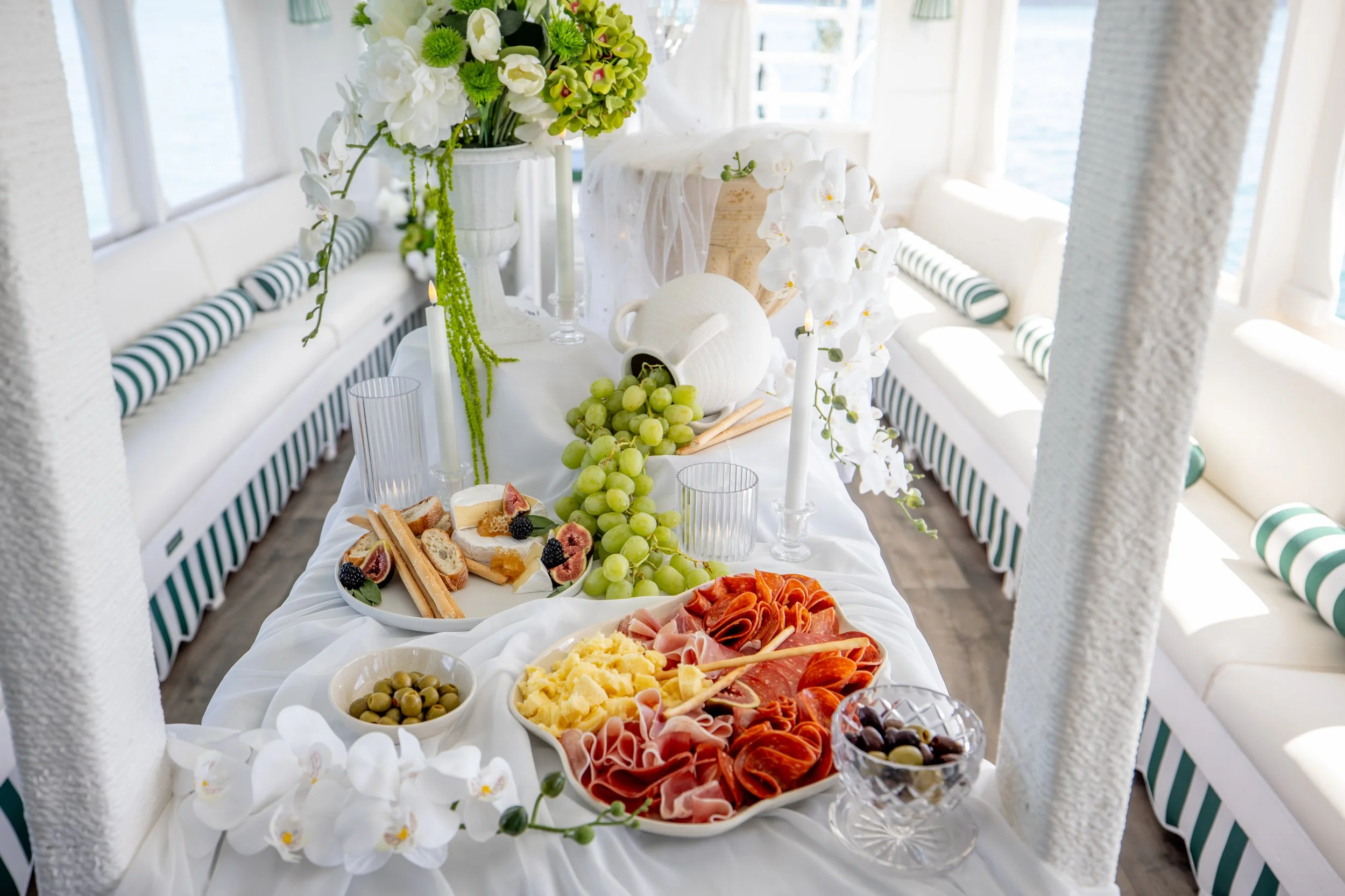 A close-up of a buffet table with various appetizers and fruits, including grapes, ham, cheese, figs, and olives, set against a white, floral-decorated backdrop with a window in the background.