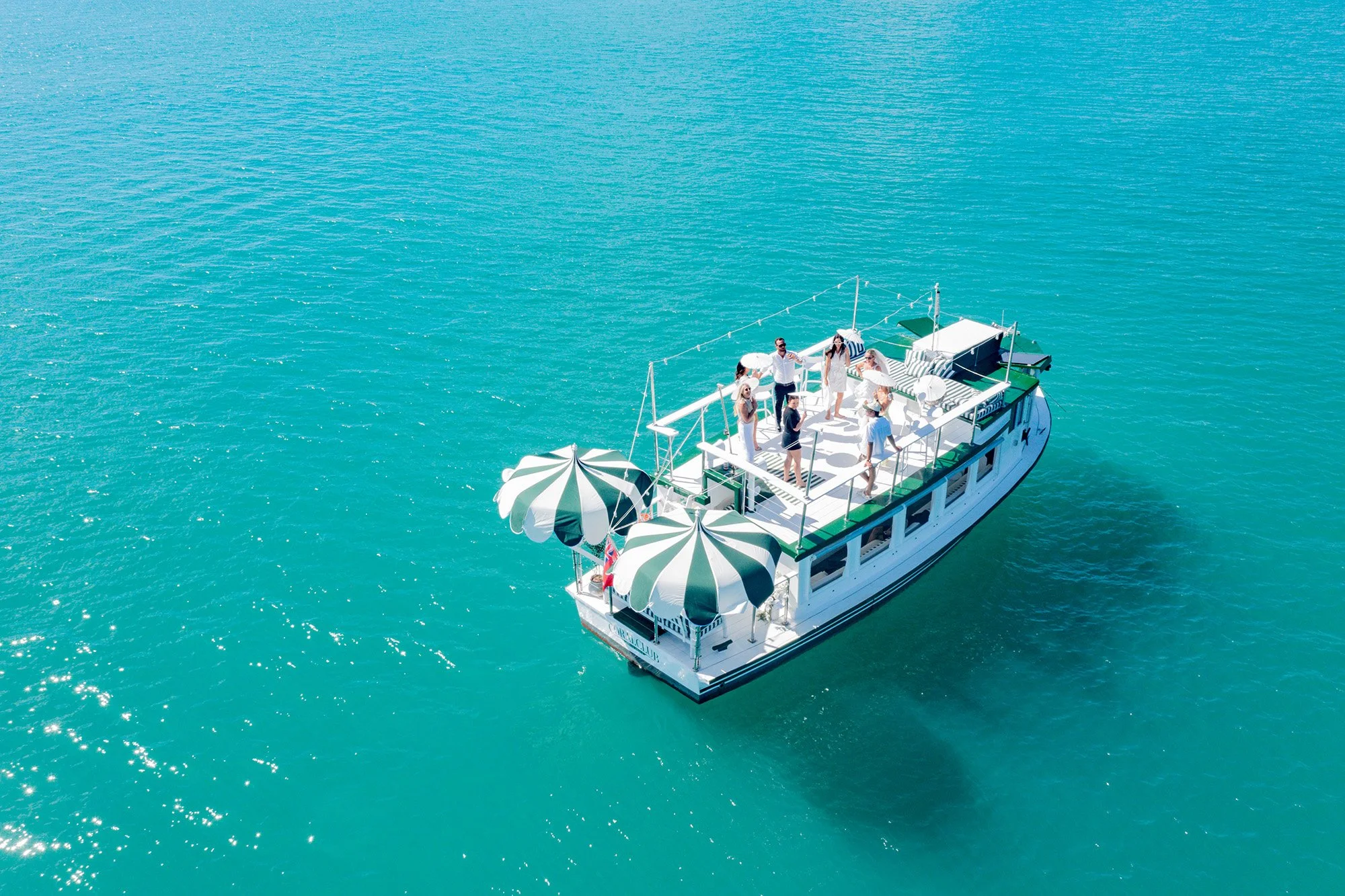 Bird's eye view of a group of people enjoying a boat cruise on a bright sunny day in the turquoise water. The boat is decorated with green and white striped umbrellas and outdoor furniture.