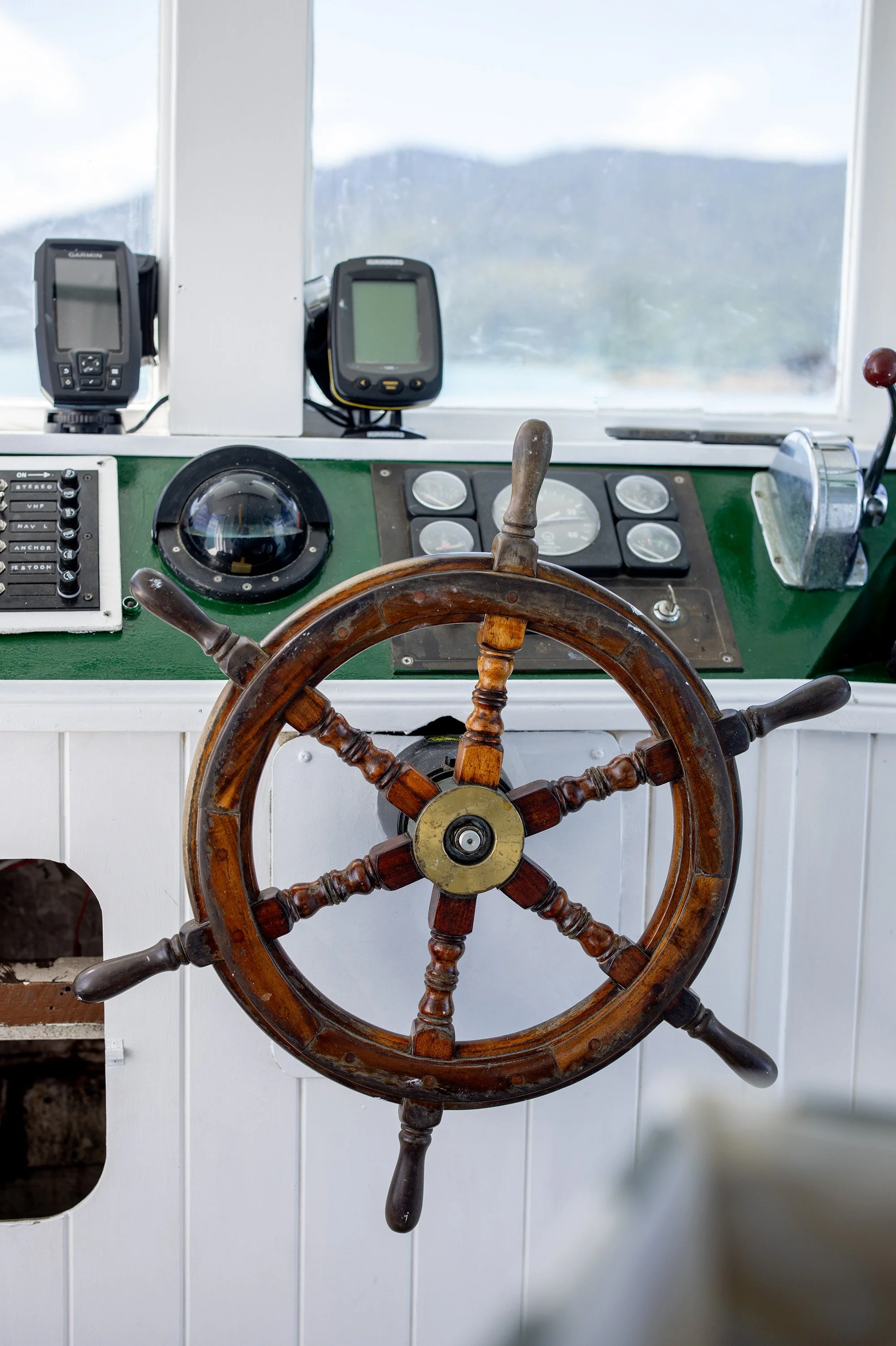 Close-up view of a steering wheel with wooden and metal parts, a ship's controls panel, and Garmin electronic navigation devices in the background on a boat's helm station.