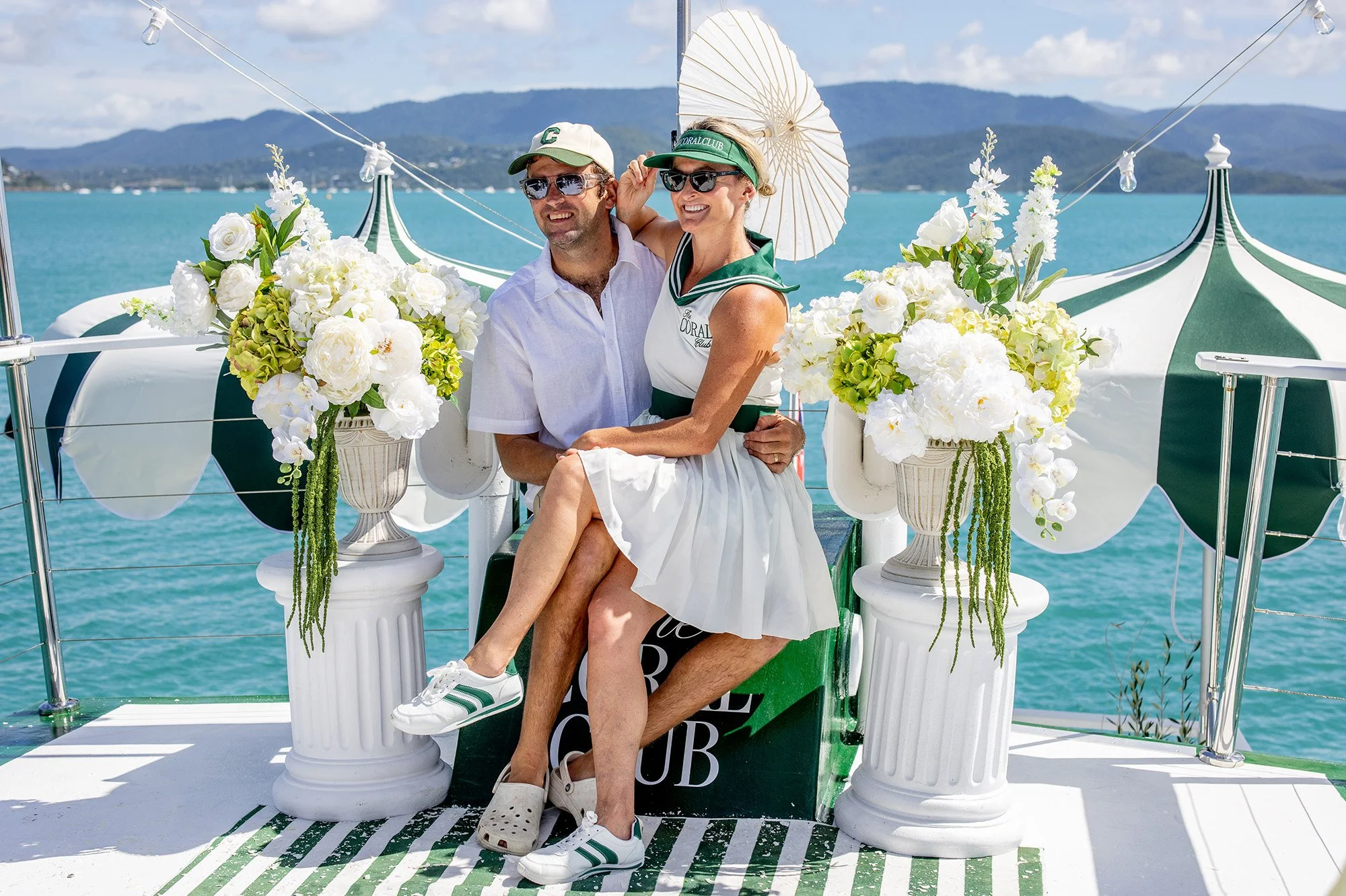 A couple sitting on a decorated platform by the water, with mountains in the background. The man is wearing a light purple shirt, sunglasses, and a white cap. The woman is wearing a white dress, sunglasses, a green visor, and is holding a white parasol. They are surrounded by white floral arrangements and the platform is adorned with white columns and decorative elements. The scene appears festive and scenic, likely at a boat or water-themed event.