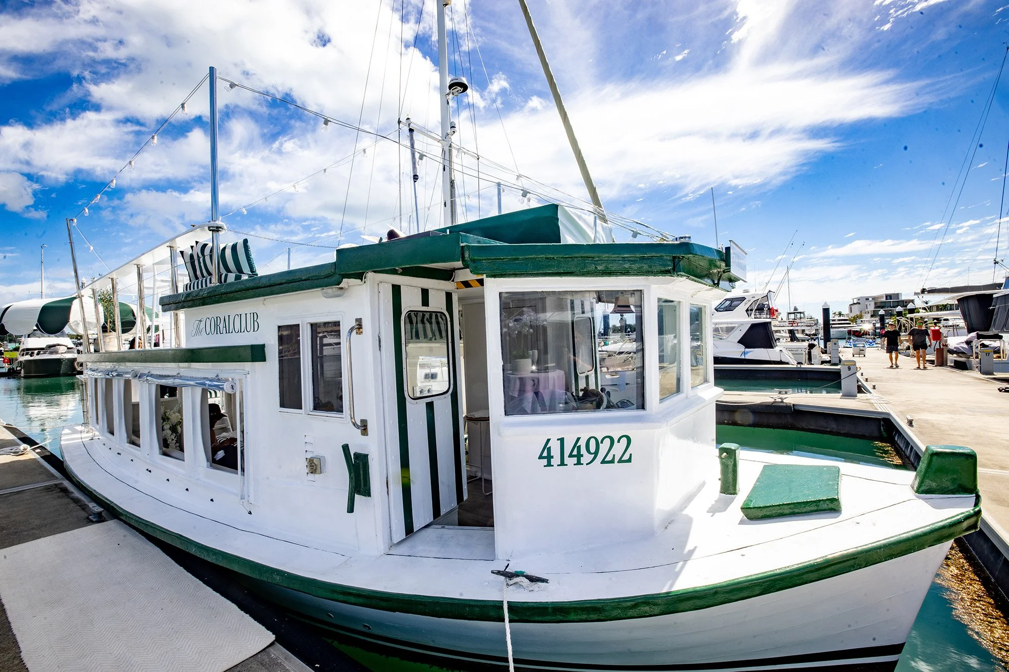 A white and green boat docked at a marina, with the name 'The Coral Club' and the number '414922' visible on the boat. Other boats and people walking are visible in the background under a partly cloudy sky.