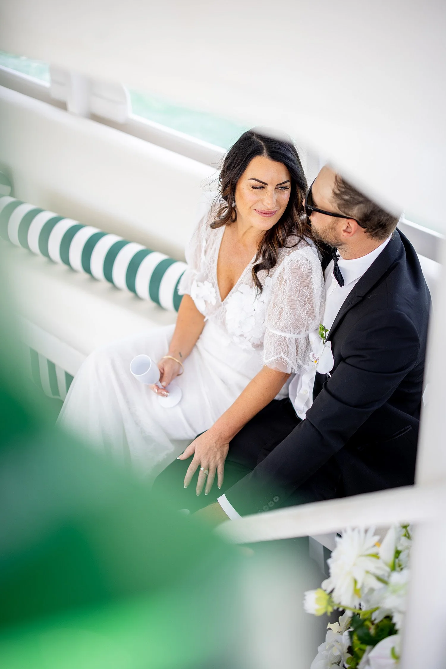A bride and groom sitting close together on a white bench with a green-striped cushion, facing each other and sharing a happy moment at their wedding. The bride is holding a white wedding program and wearing a white lace wedding dress. The groom is dressed in a black tuxedo with a white shirt and black bow tie. Floral decorations and white flowers are seen in the foreground.