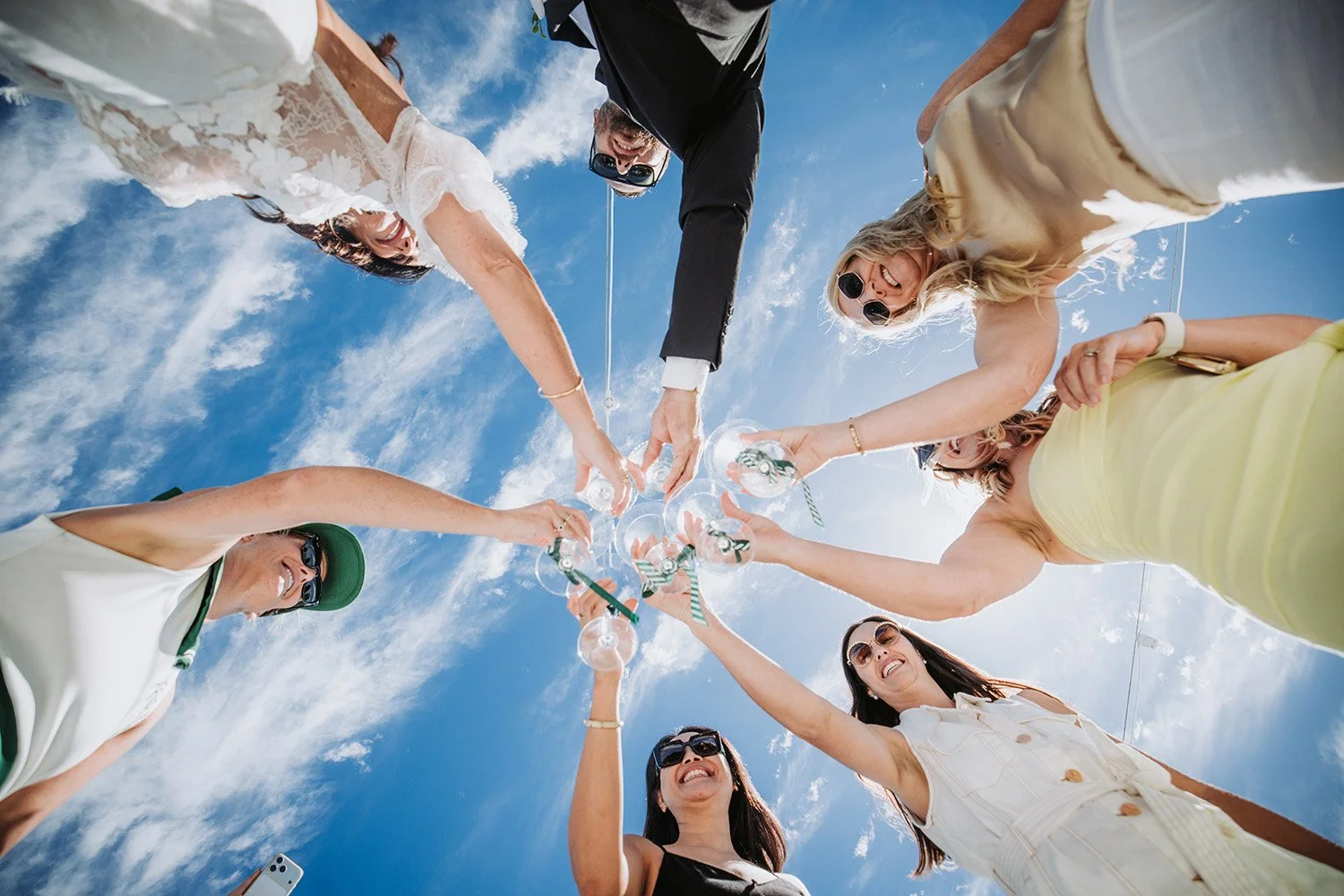 Group of friends at a beach, standing in a circle, clinking glasses filled with drinks, smiling, and enjoying sunshine with a blue sky and clouds in the background.