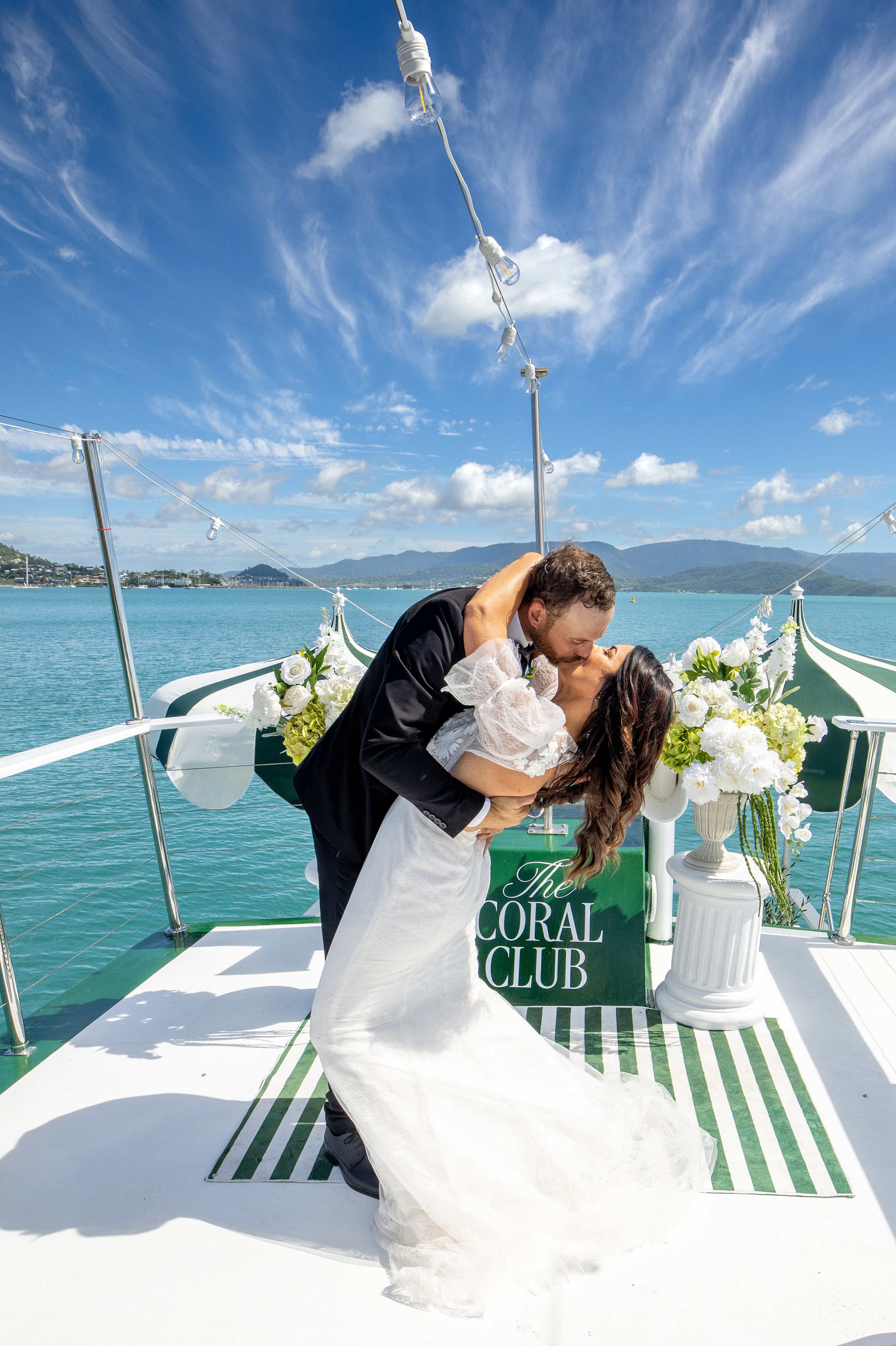 A newlywed couple kissing on a boat decorated with white flowers and a sign for The Coral Club, with water, mountains, and a blue sky with clouds in the background.