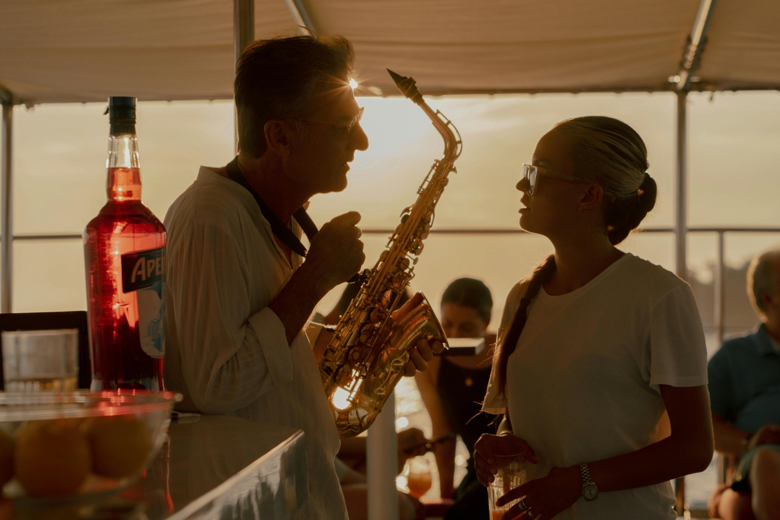 A man and woman standing and interacting at a concession stand or bar during sunset, with the man holding a saxophone and the woman holding a plastic cup.
