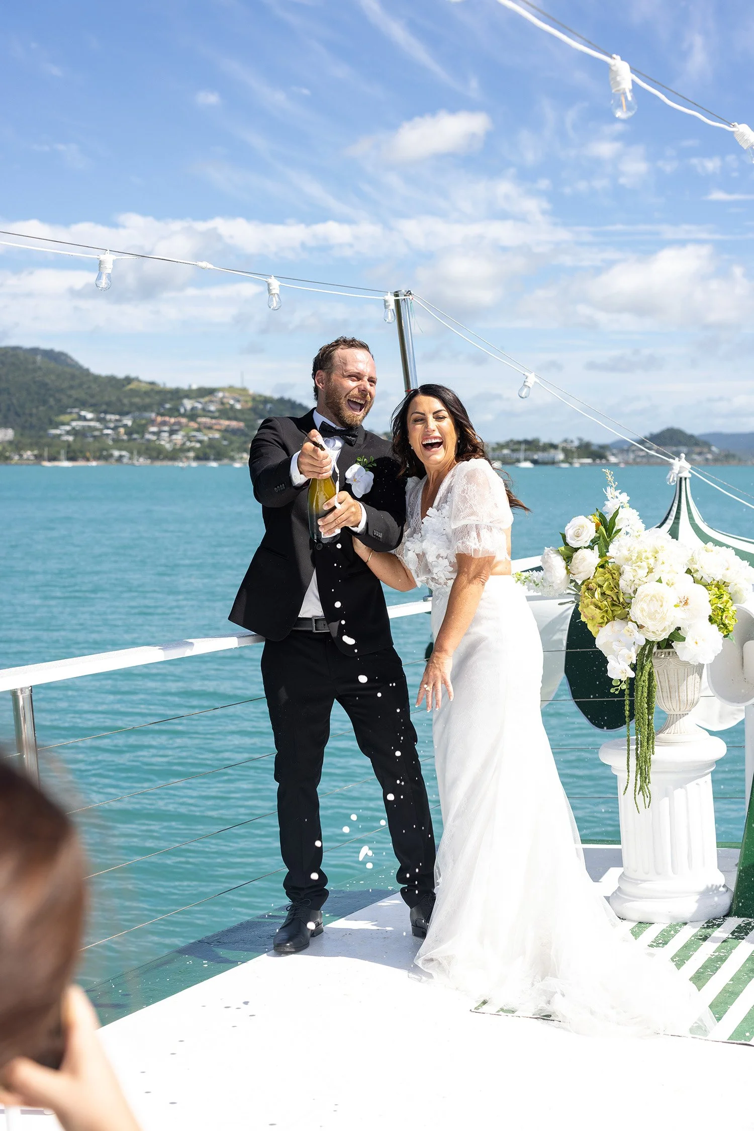A newlywed couple celebrating on a boat, with the groom spraying champagne and the bride laughing, against a backdrop of water and mountains, under a sunny sky with string lights.