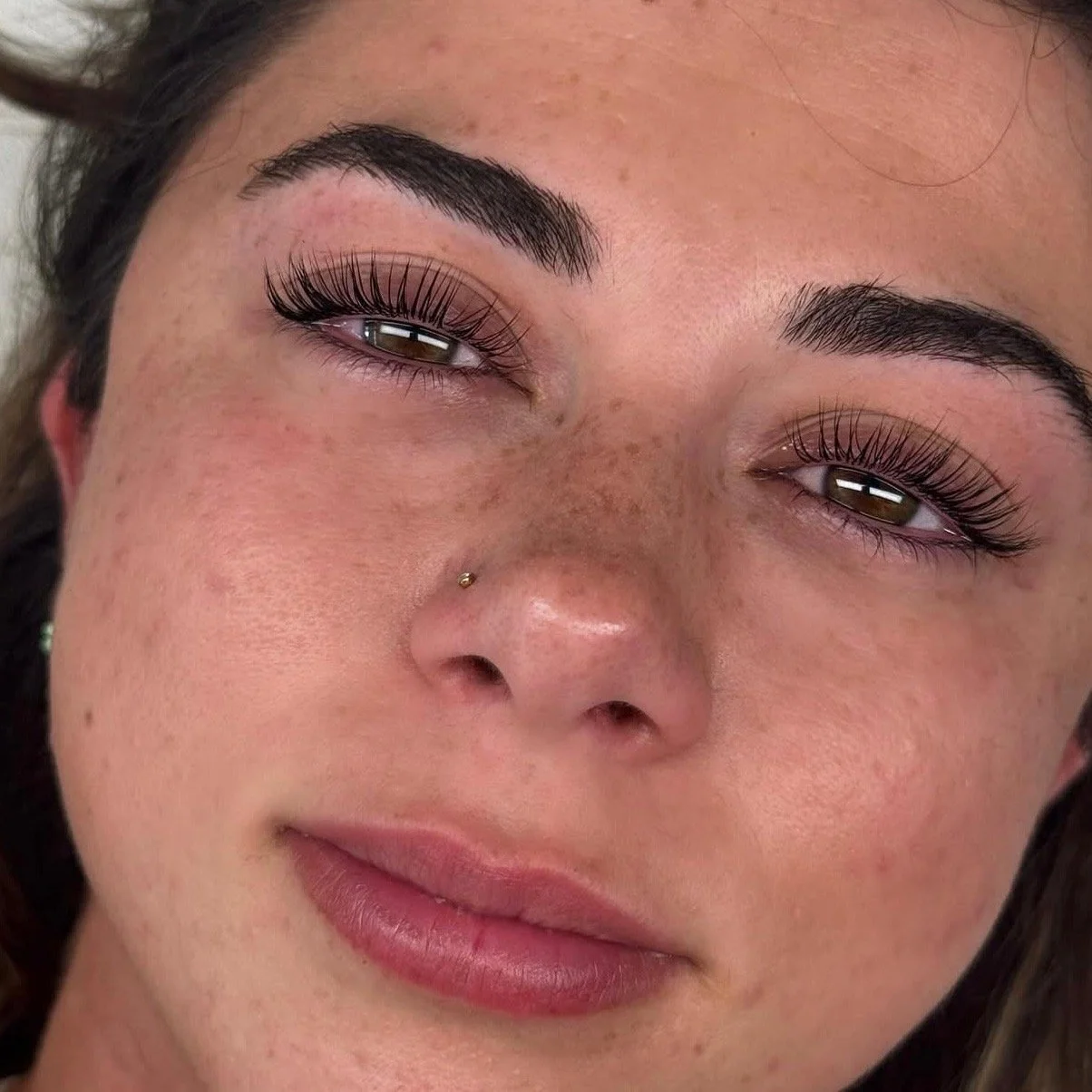 Close-up of a young woman's face with natural skin, well-groomed eyebrows, long false eyelashes, brown eyes, a small nose piercing, and light pink lips, with her eyes half-closed.
