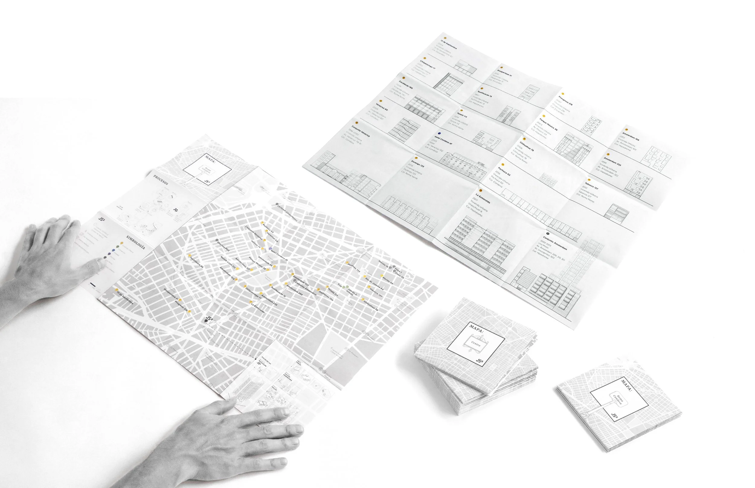 Hands arrange architectural plans, maps, and booklets on a white surface, including a city map with marked locations. The plans feature building elevations and diagrams.