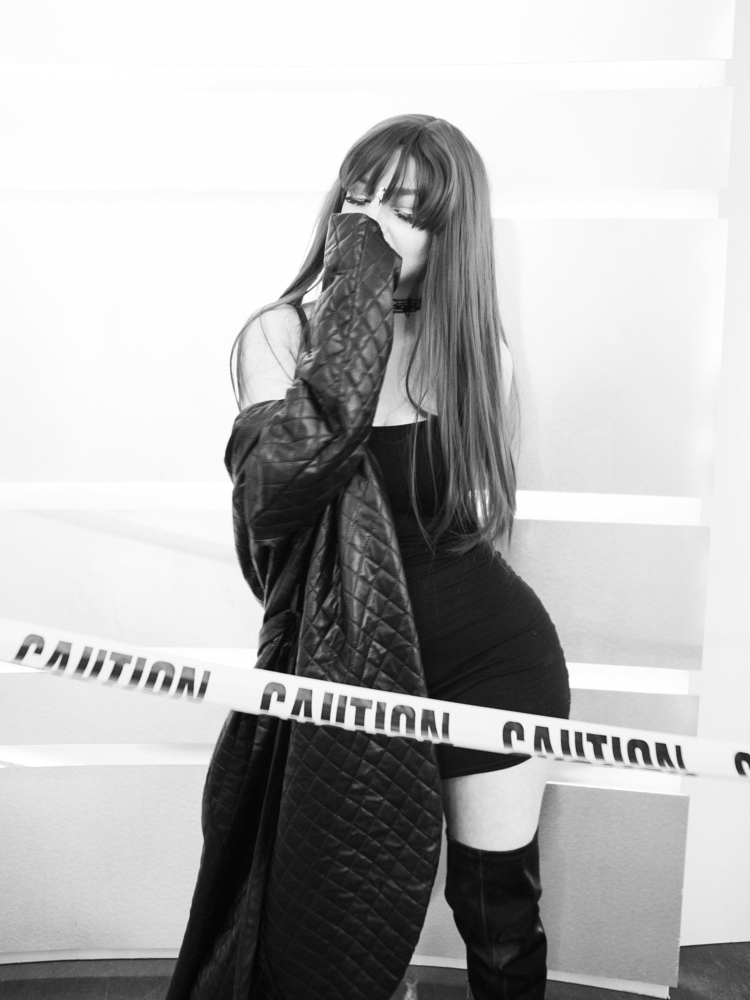A woman with long hair and bangs is holding a quilted jacket near her face, standing behind caution tape in a black and white photo.