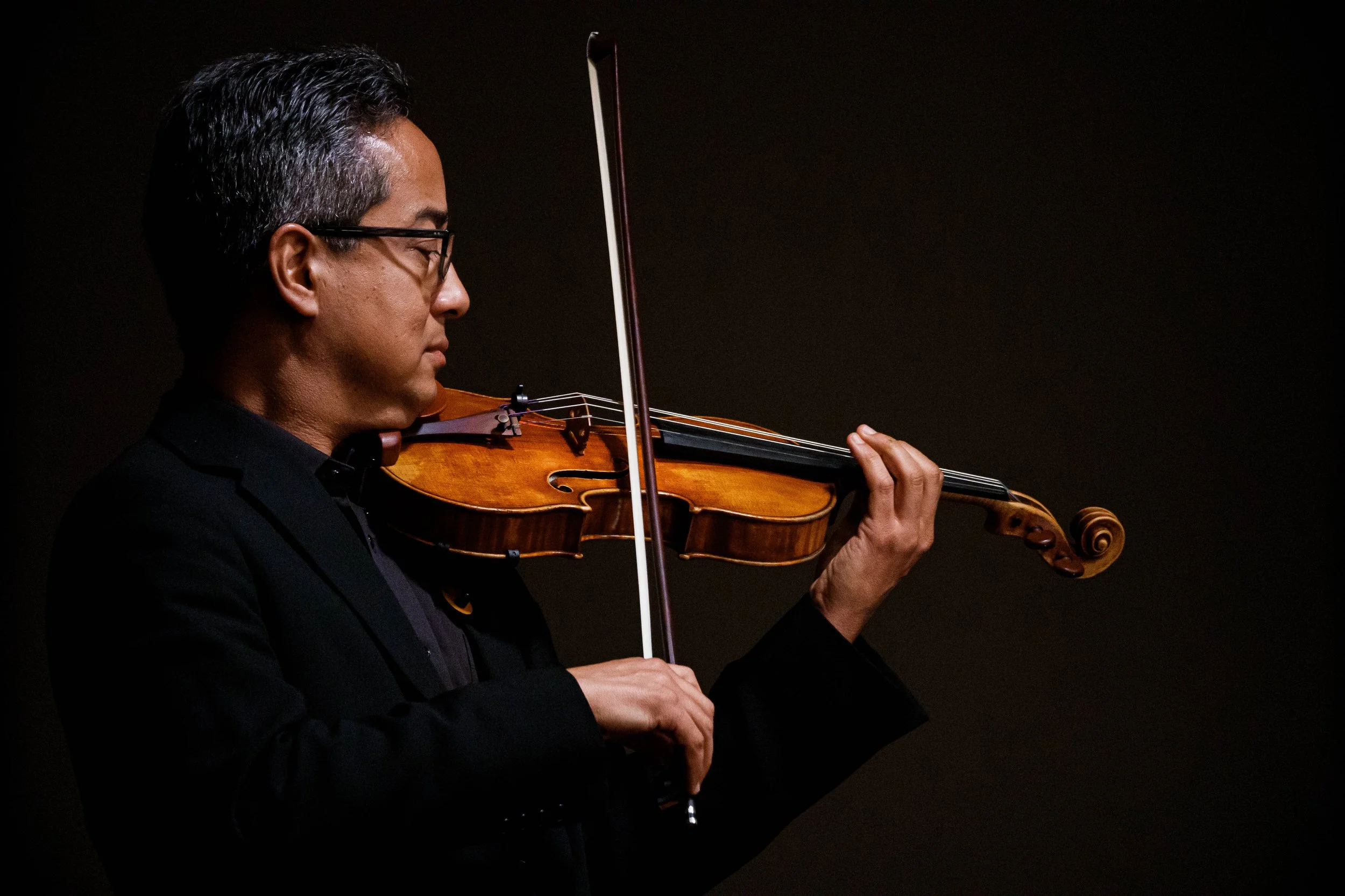 A man playing a violin against a dark background, wearing glasses and a black suit.
