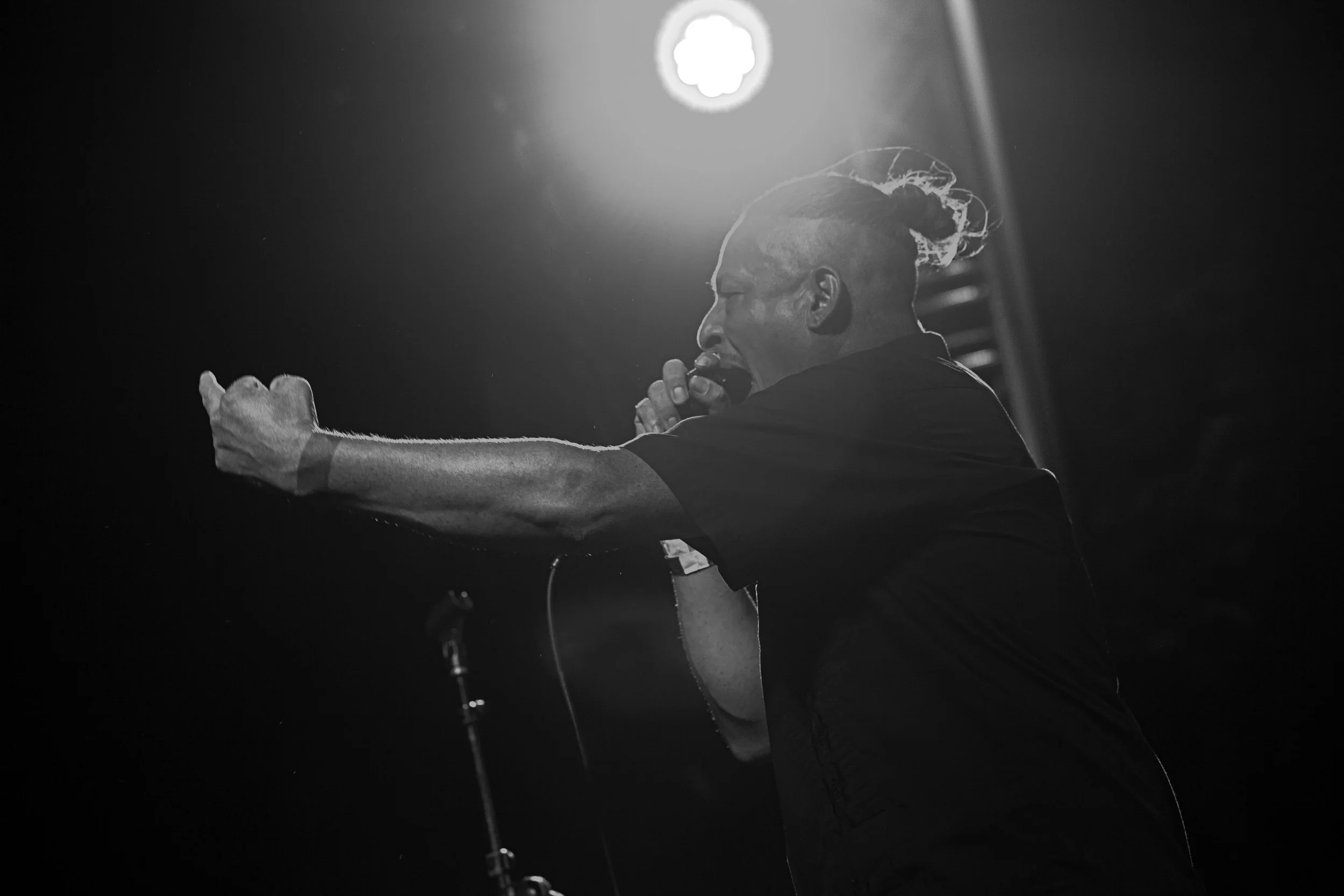 A person with a mohawk hairstyle passionately singing into a microphone on stage, with a bright overhead stage light shining behind them in a black and white photo.