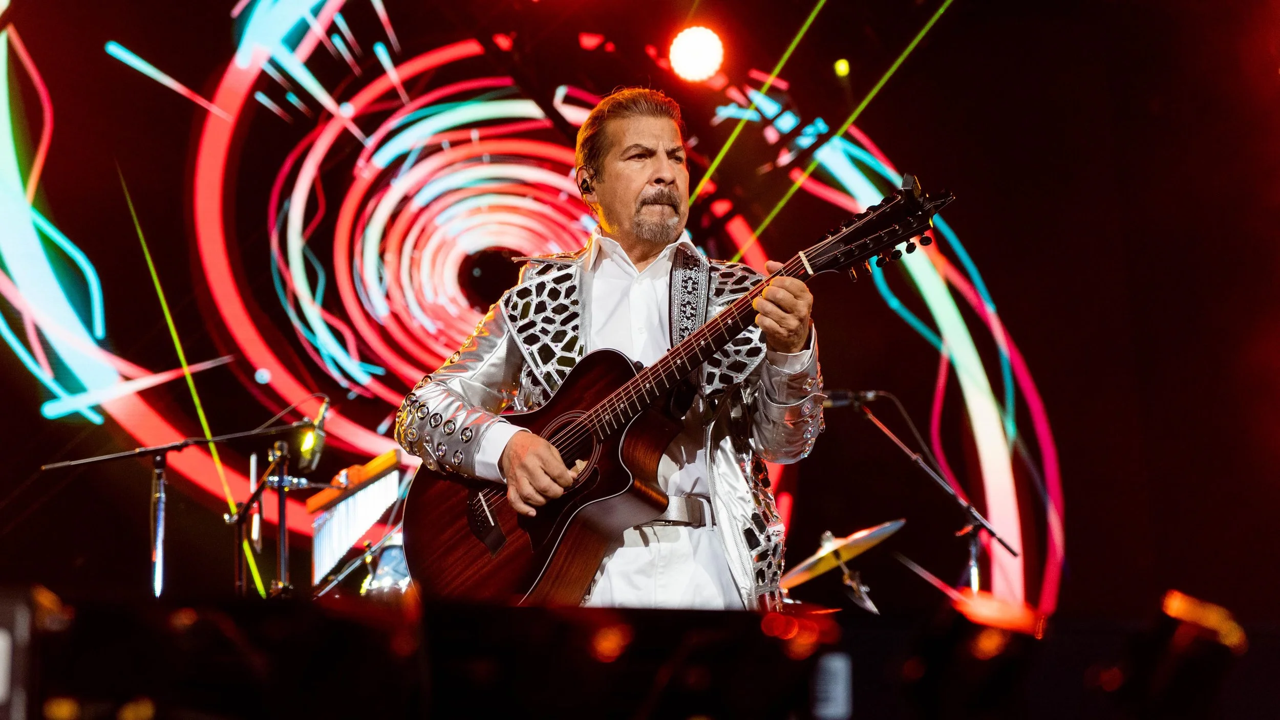 Man playing acoustic guitar on stage with colorful spiral light display behind him.