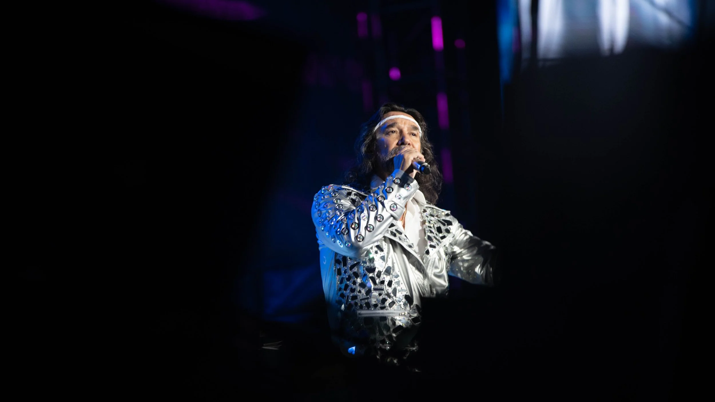 Marco Antonio Solis, a male performer with long hair, wearing a shiny silver jacket with decorative elements, singing into a microphone on stage with dark background and purple stage lights.