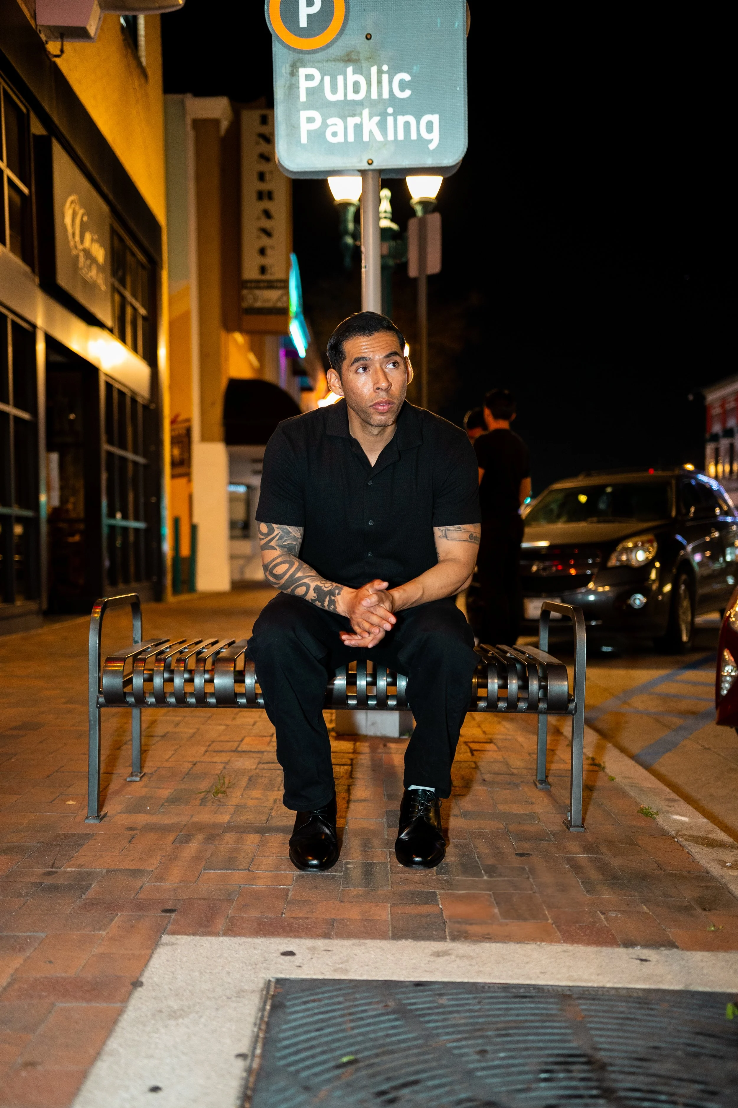 A man sits on a metal bench on a city sidewalk at night, wearing a black shirt and black pants, with tattoos on his arms, under streetlights, with cars parked along the street and a sign indicating public parking.