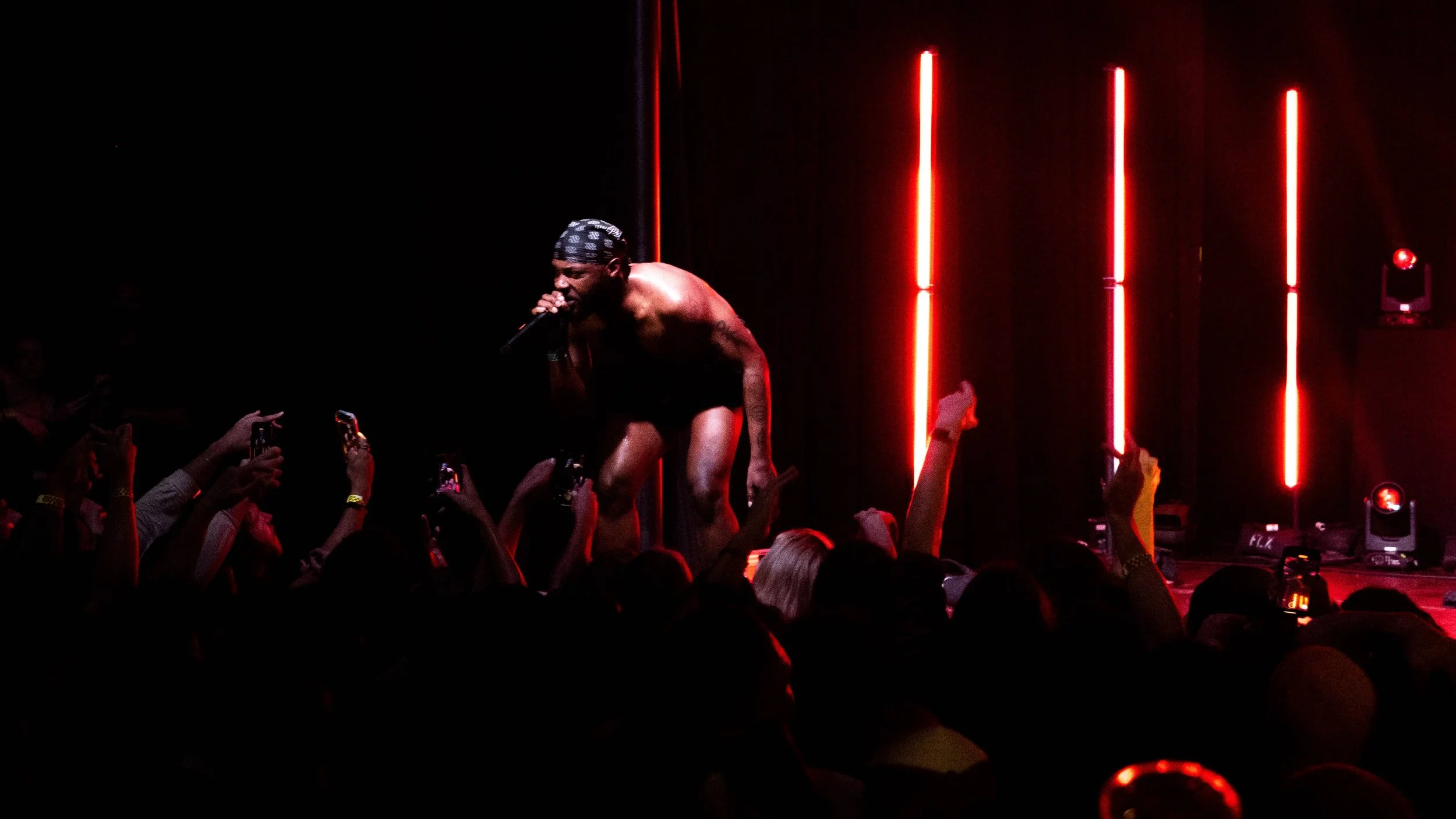 JPEGMAFIA,  shirtless male performer with a bandana on his head crouching and singing into a microphone on stage, surrounded by an audience taking photos and raising their hands, with red vertical stage lights in the background.