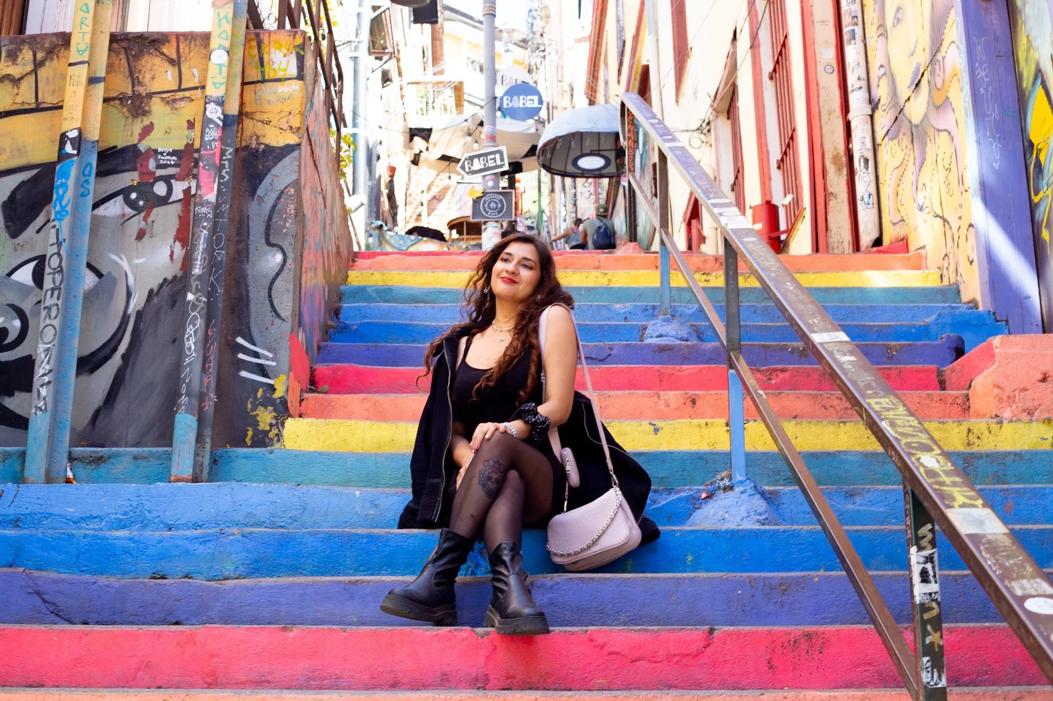 A young woman with long wavy hair sitting on colorful painted stairs in an urban area with graffiti, smiling at the camera.