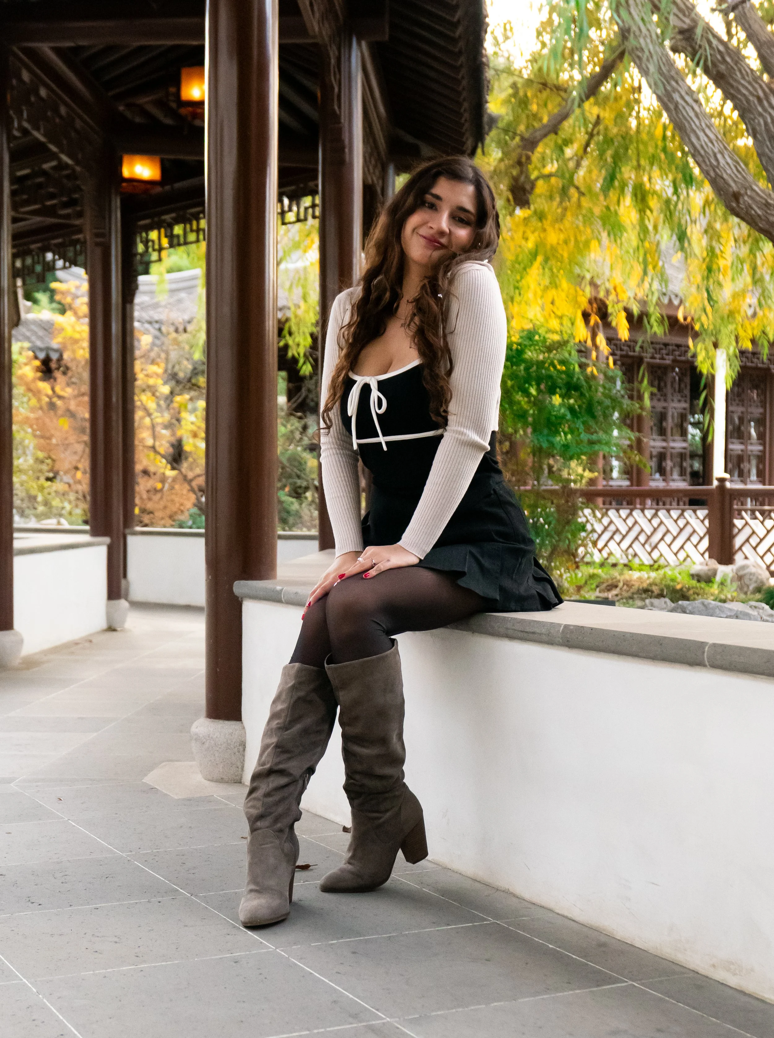 A young woman with long curly hair, smiling, sitting on a ledge outside a traditional Asian-style building with wooden pillars, surrounded by green and yellow autumn trees.
