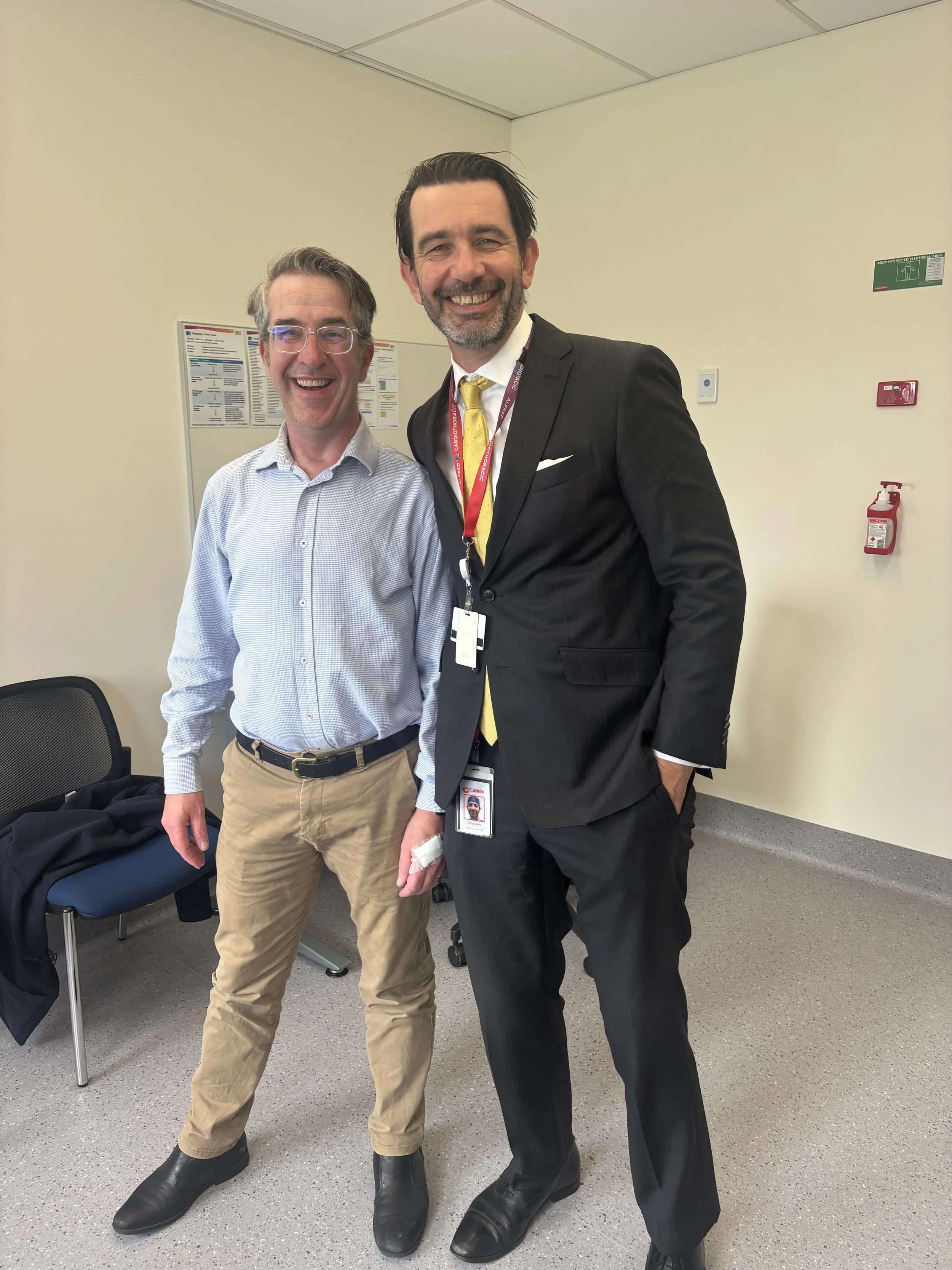 Two men standing together, smiling in an office or conference room, one wearing a suit and the other in a dress shirt and khakis.