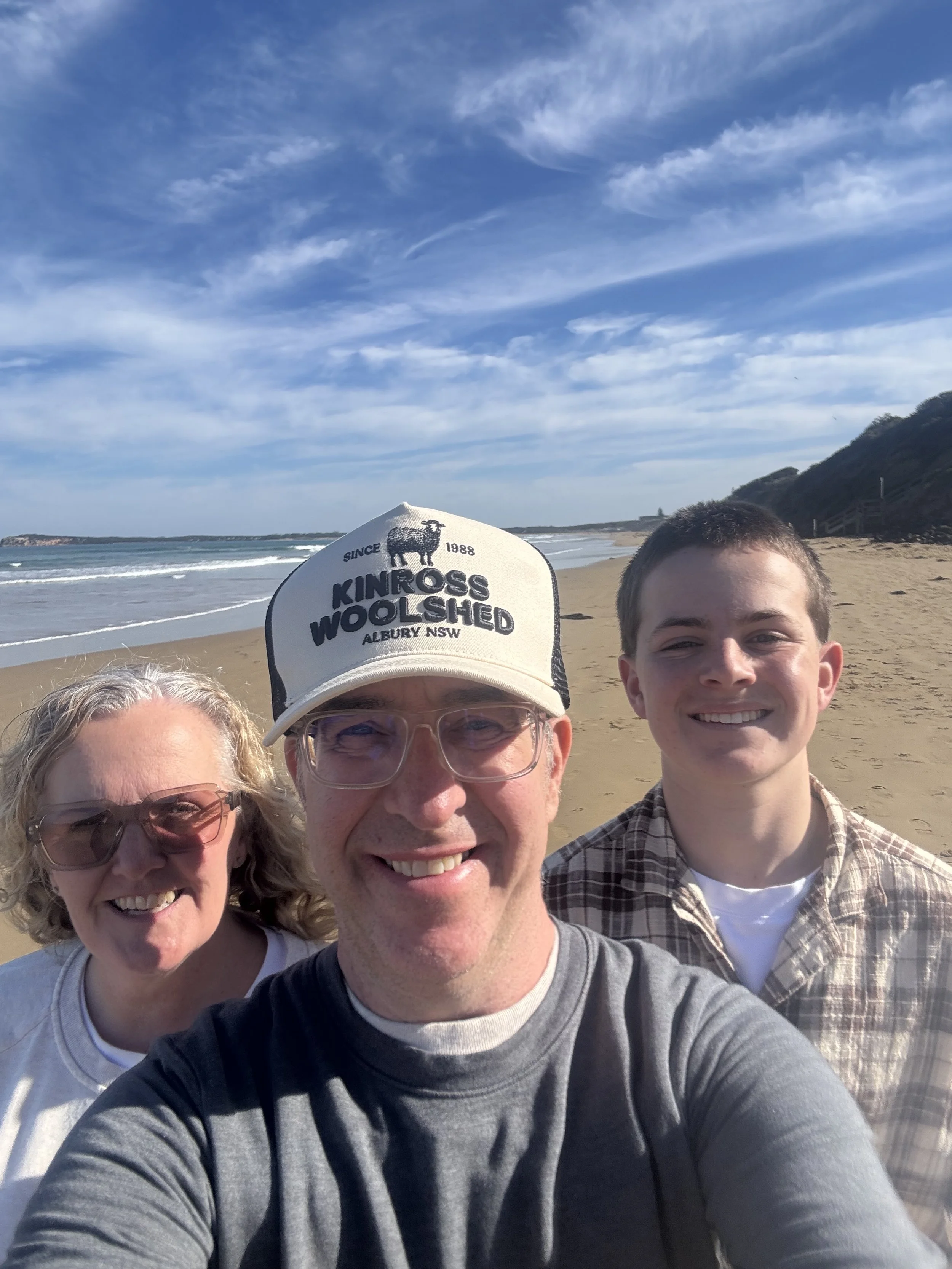 Family taking a selfie on the beach with blue sky and waves in the background.