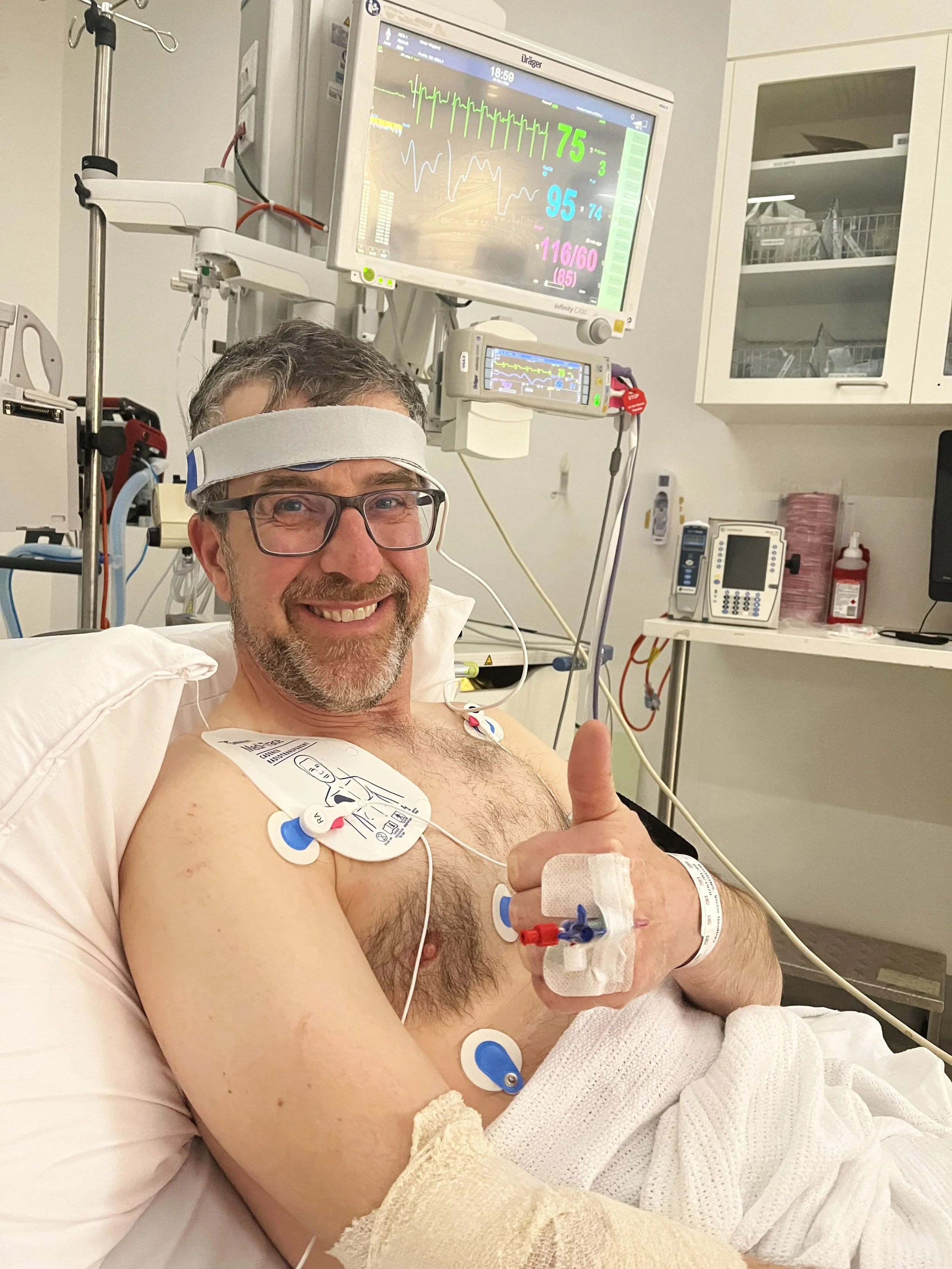 A smiling man in a hospital bed giving a thumbs-up gesture with his right hand. He is wearing glasses and has medical monitoring devices attached to his chest and arm. He is wearing a headband with a sensor. Behind him, medical equipment and monitors