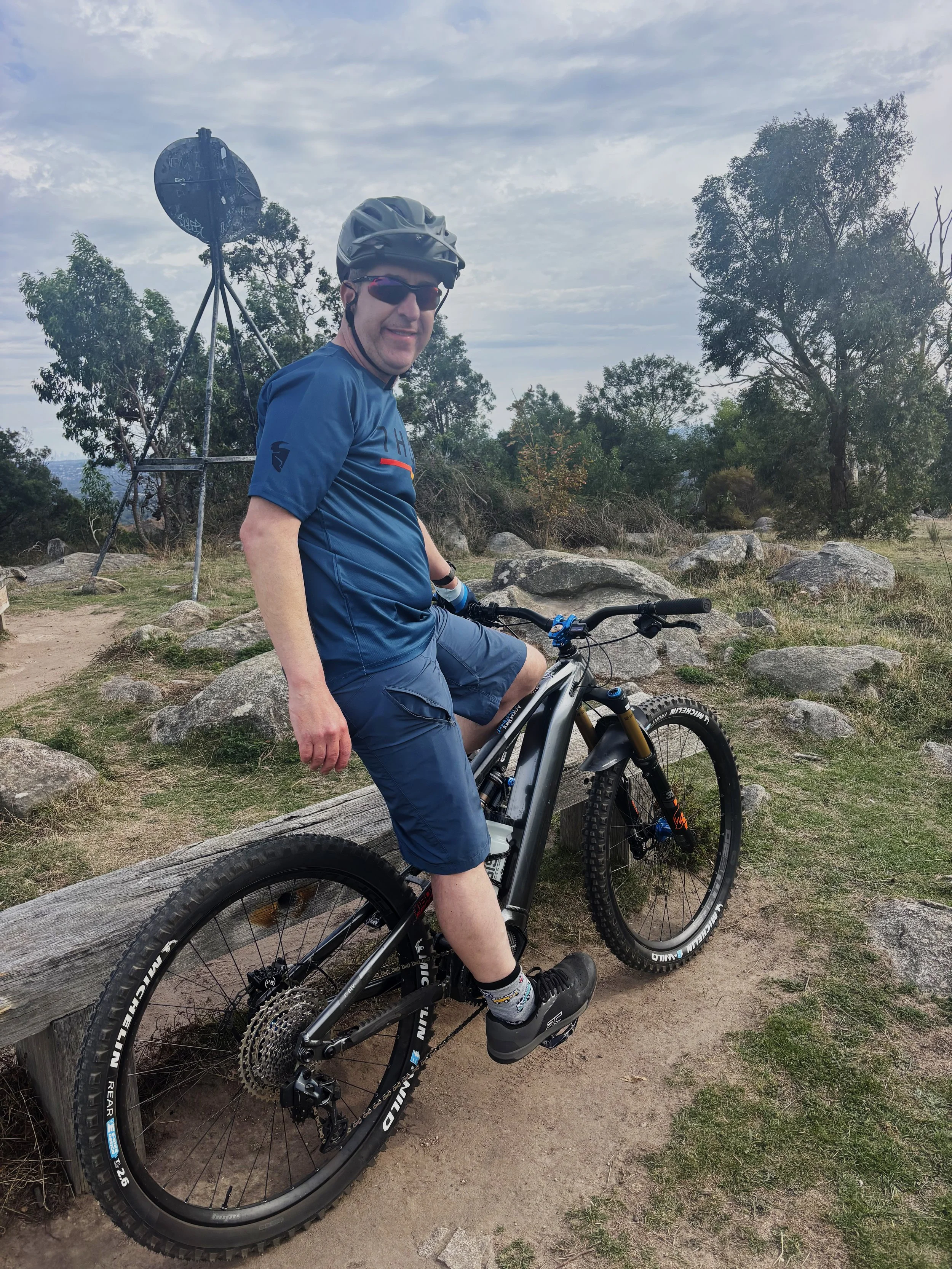 Man wearing a helmet and sunglasses riding a mountain bike on a dirt trail surrounded by rocks and trees.