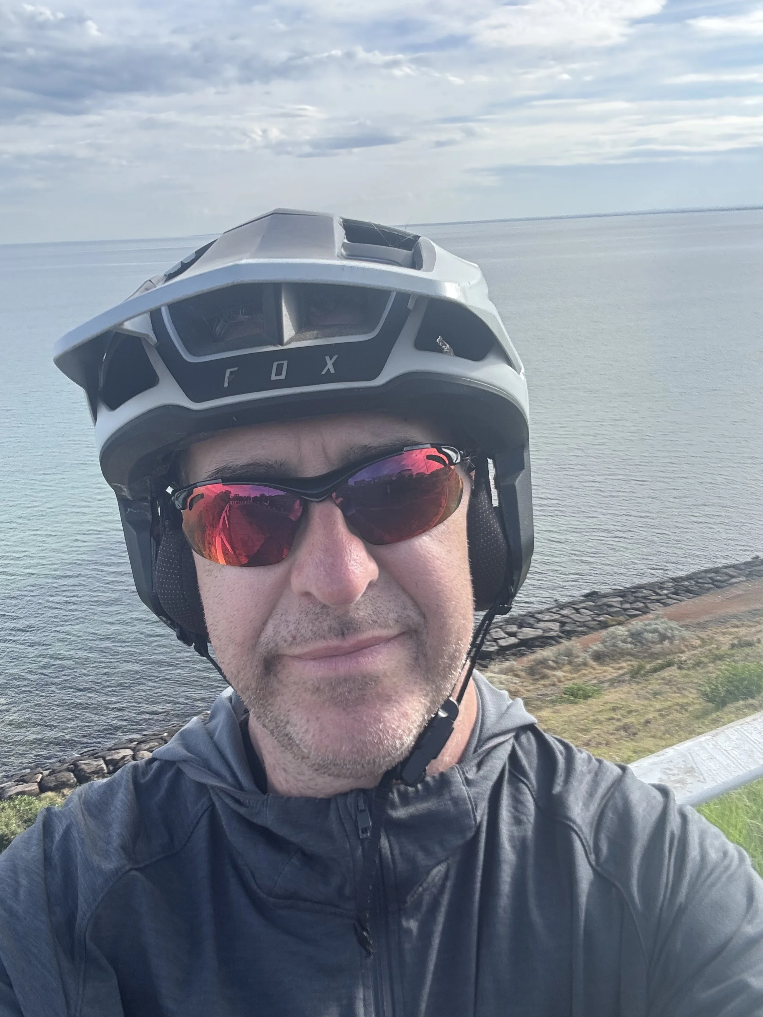 Man wearing a helmet and sunglasses taking a selfie near a body of water with a sky filled with clouds.