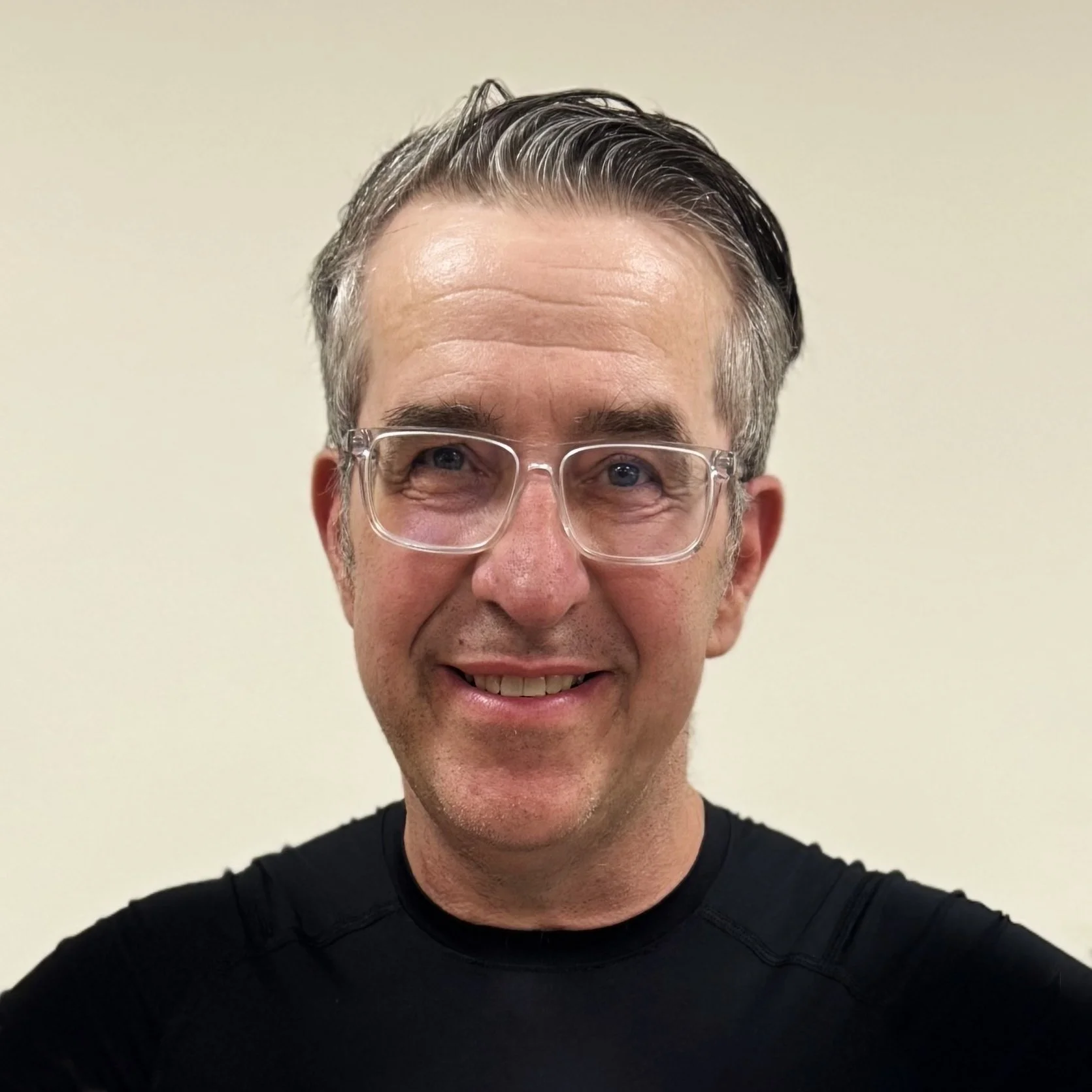 A smiling middle-aged man with gray hair, wearing clear glasses and a black athletic shirt, standing against a plain light-colored background.