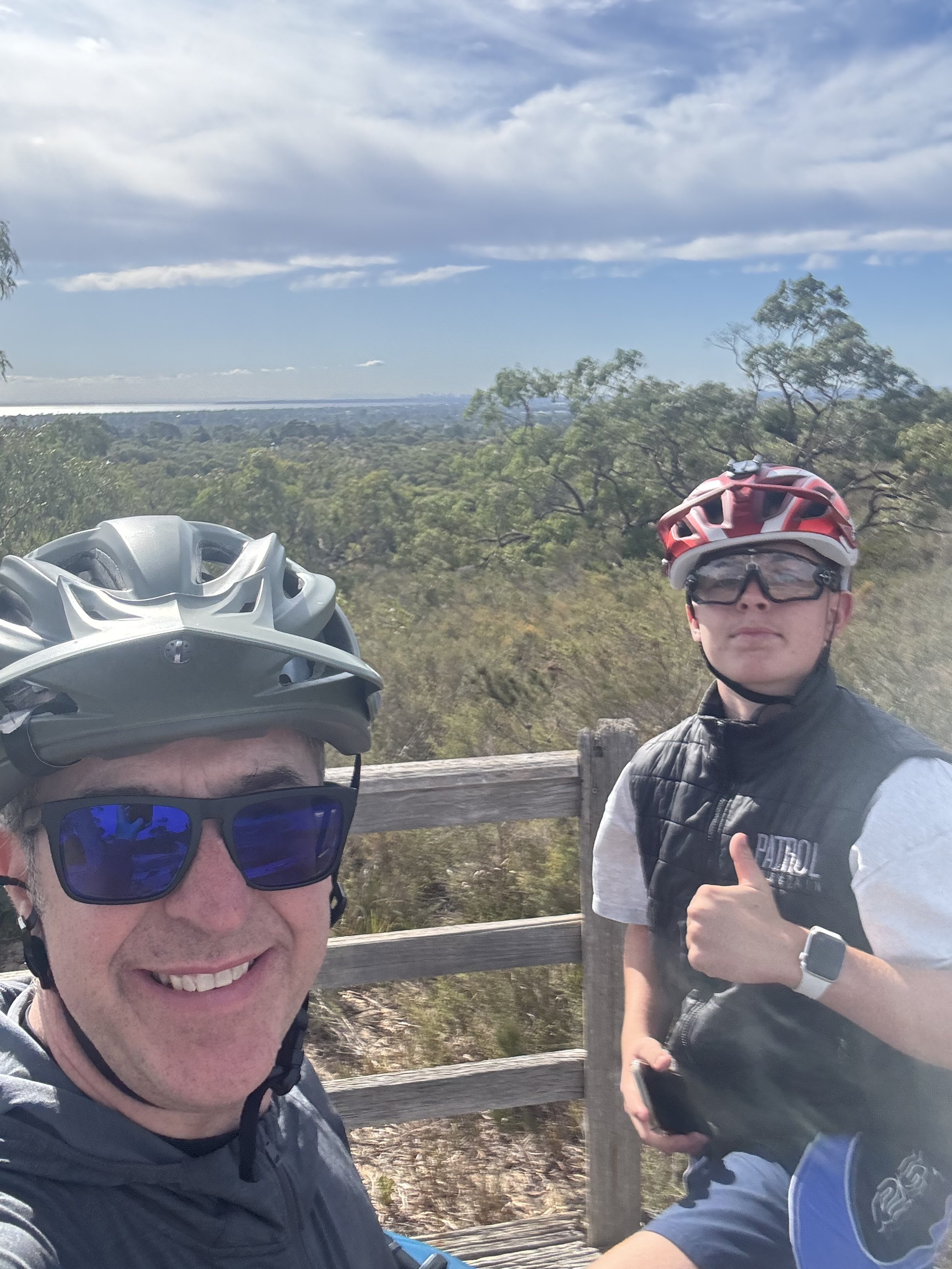 Two cyclists wearing helmets and sunglasses standing on a wooden fence, with a scenic landscape of trees and a cloudy sky in the background.