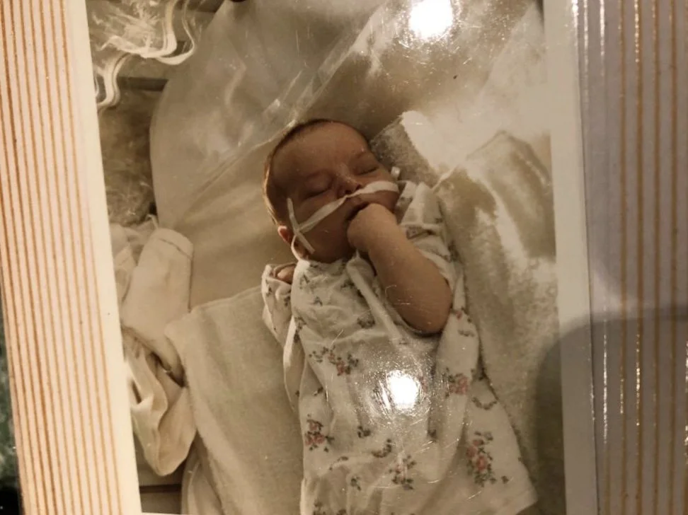 A baby sleeping in an incubator with a nasal tube and an oxygen mask, dressed in a floral onesie, in a hospital setting.