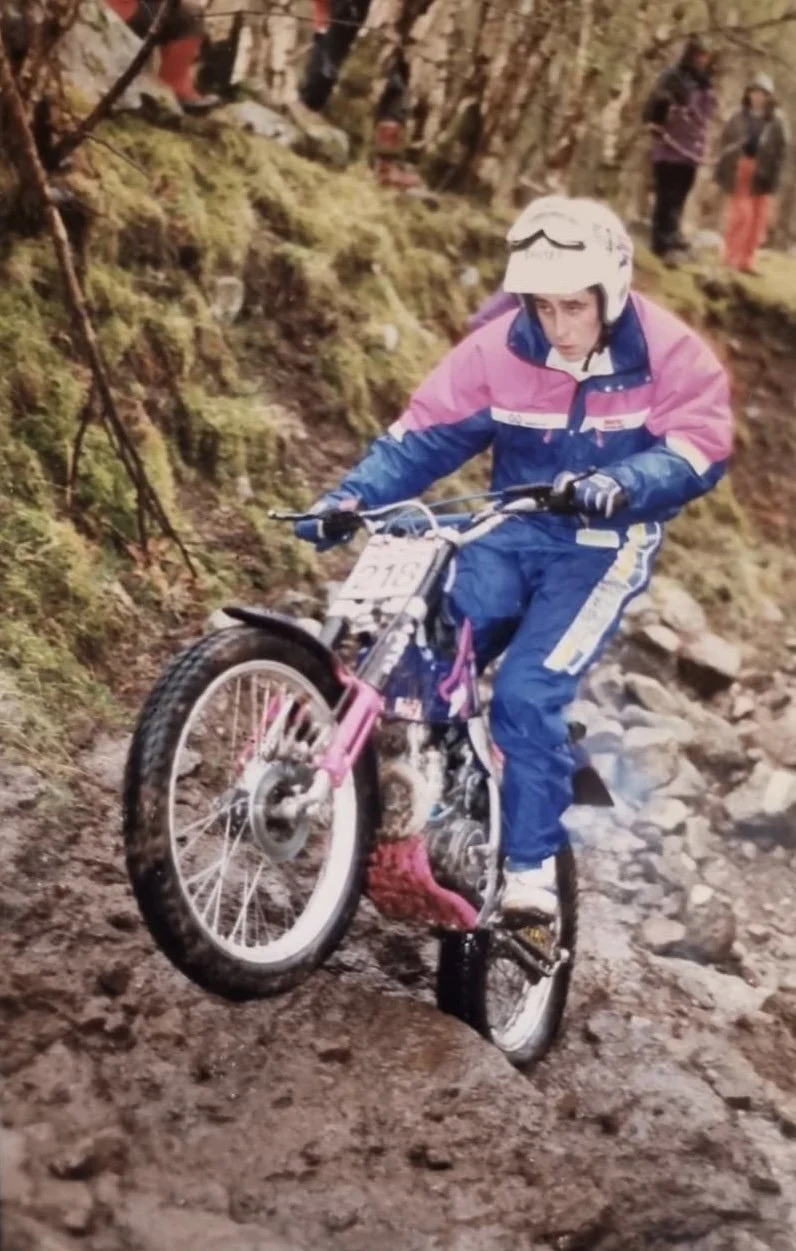 A woman riding a motorized trial bike on a rocky trail in the woods, wearing a helmet and colorful outdoor clothing.