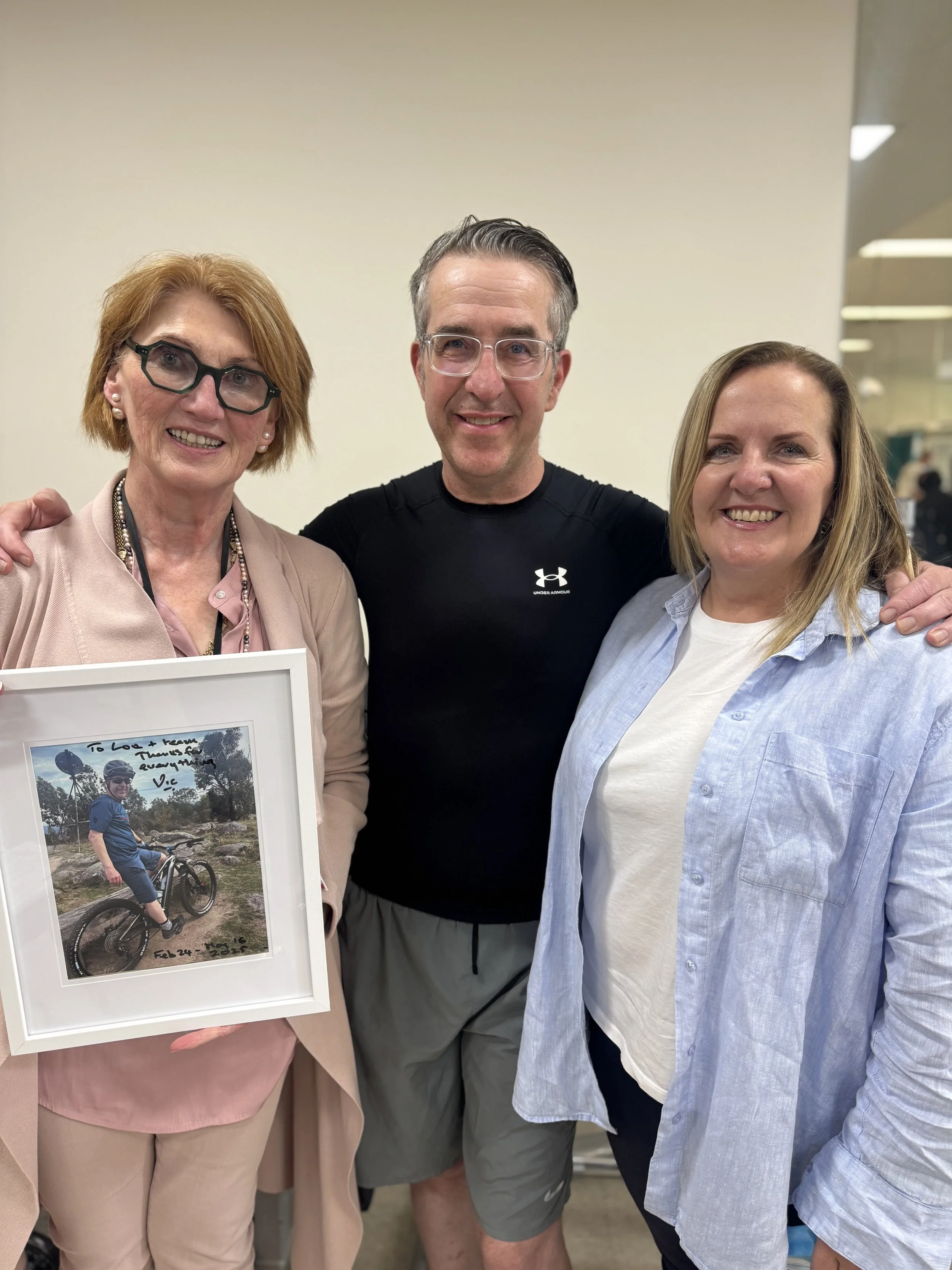 Three adults, two women and one man, smiling and posing in an indoor setting. The woman on the left holds a framed photo of a person riding a bike outdoors, with handwritten notes and dates on the photo.