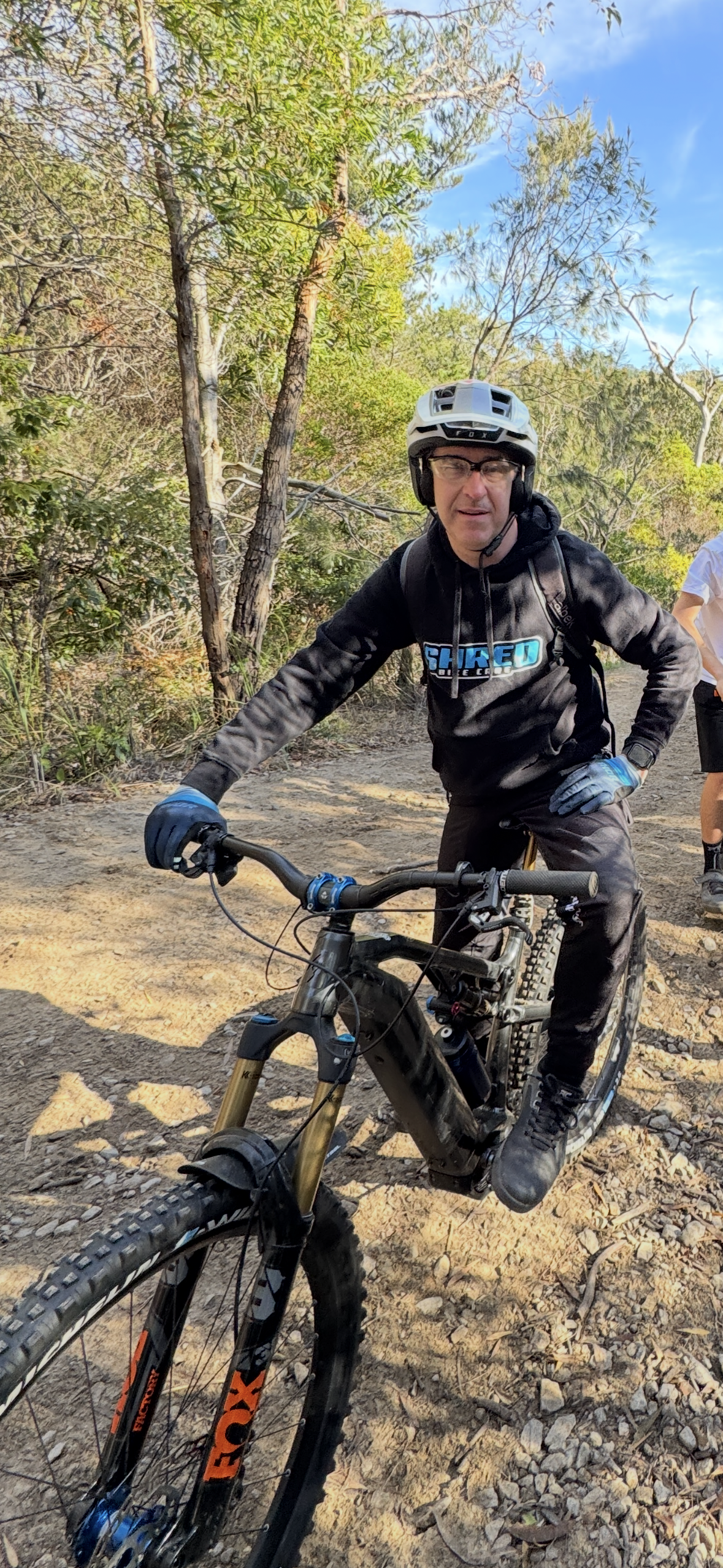 A man wearing a white helmet, glasses, a black hoodie with a logo, and gloves riding a mountain bike on a rocky trail through a wooded area.