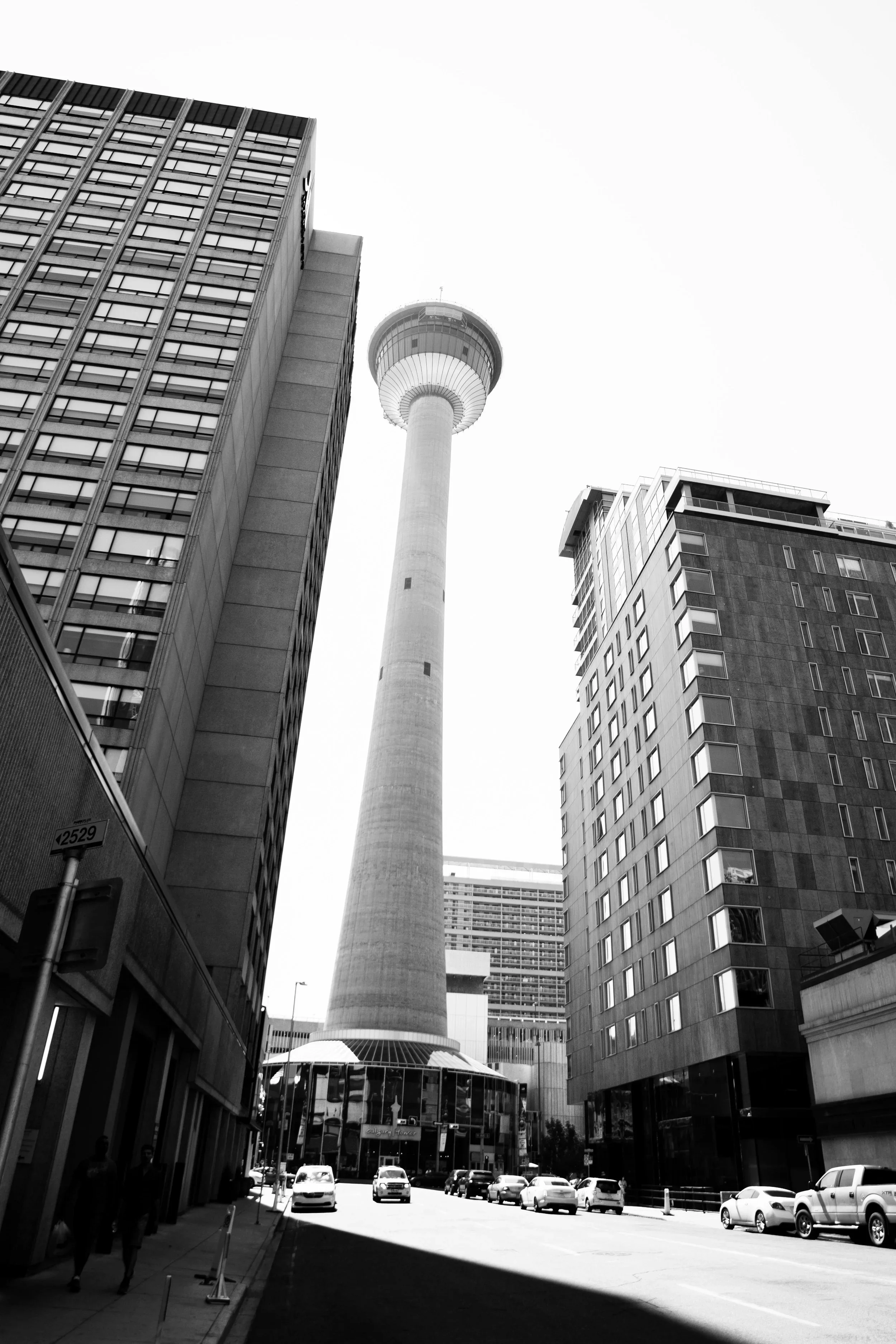 Black and white photo of an urban city street view featuring a tall communications tower surrounded by modern high-rise buildings, with cars parked and walking pedestrians.