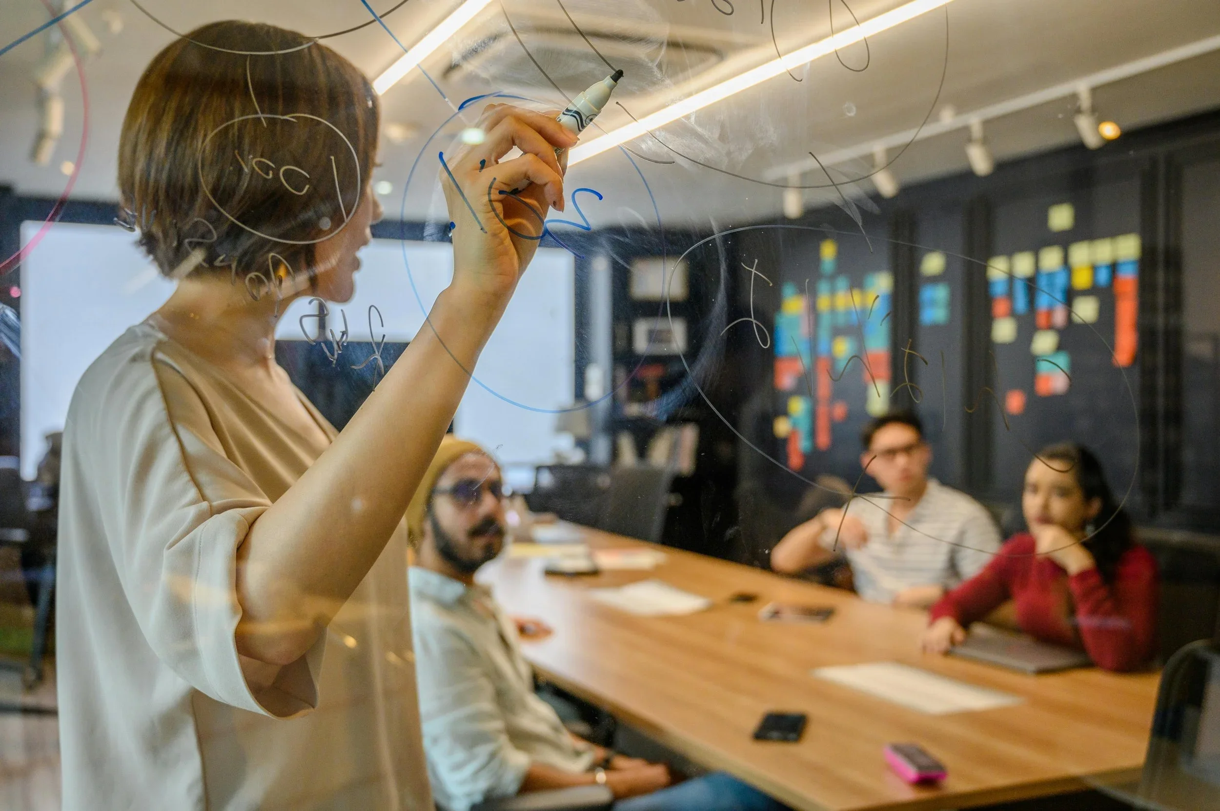 A woman writing on a glass wall with a marker while three colleagues sit at a conference table, listening.